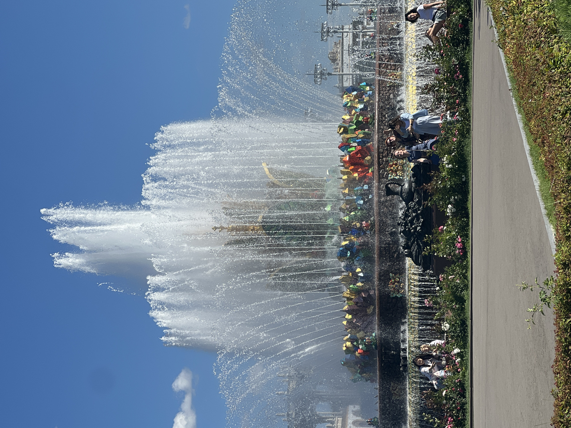 The image shows the Stone Flower fountain in Moscow, Russia, located in VDNKh park. The fountain is a large, multi-tiered structure with water jets spraying upwards and outwards, creating a luminous effect against the clear blue sky. The fountain's base is adorned with colorful, crystal-like elements, and it is surrounded by lush greenery and flowers. Several people are visible in the foreground, some sitting and posing for photos while others are standing. They are dressed in casual summer clothing. The park setting suggests a pleasant day, with a paved pathway in the foreground and decorative lampposts visible in the background.