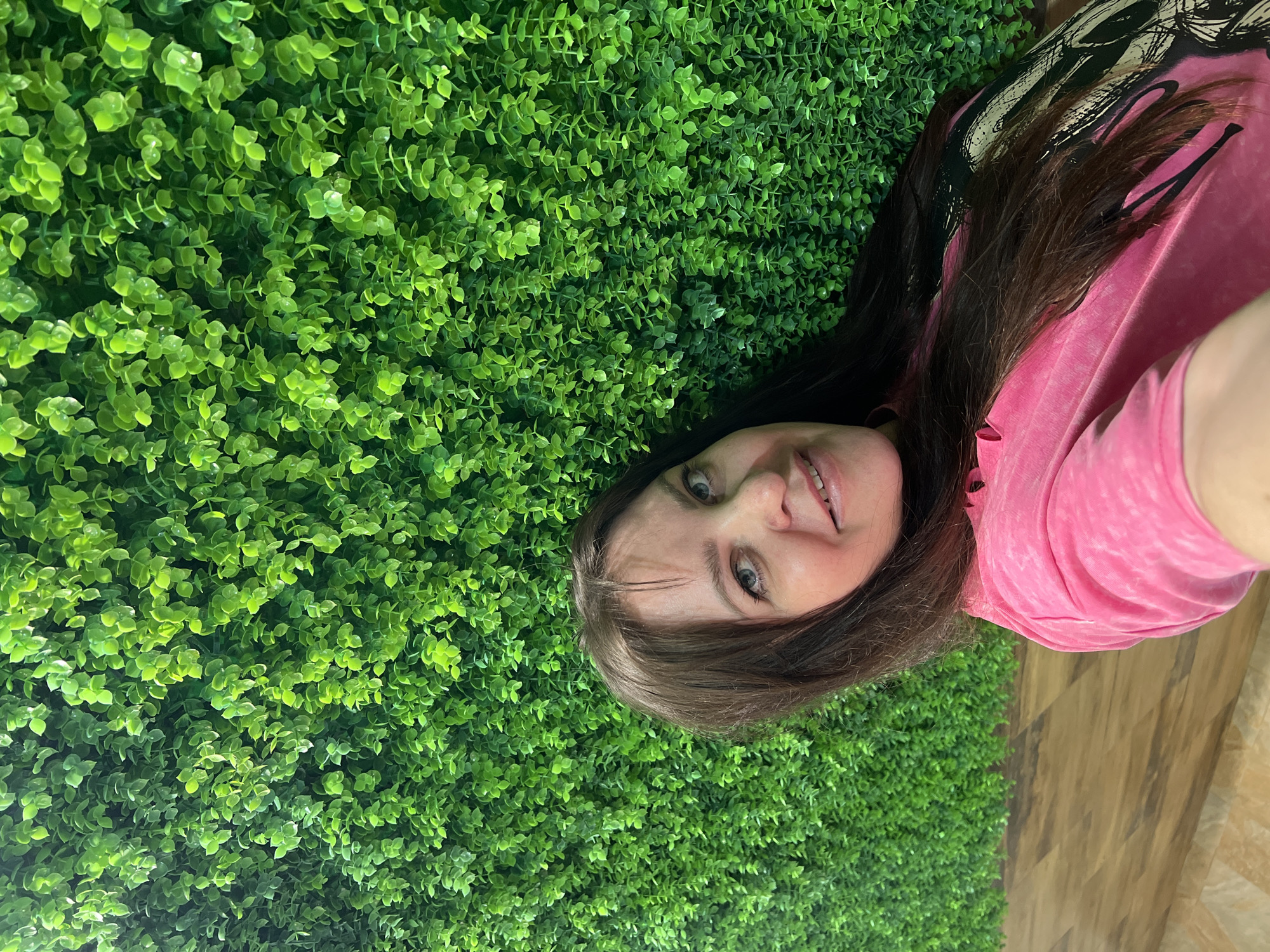 A woman lies on a green, artificial-foliage wall next to a wooden floor. She has brown hair and is wearing a pink t-shirt. The image appears to be a selfie. The location is Chaikovsky, Russia.