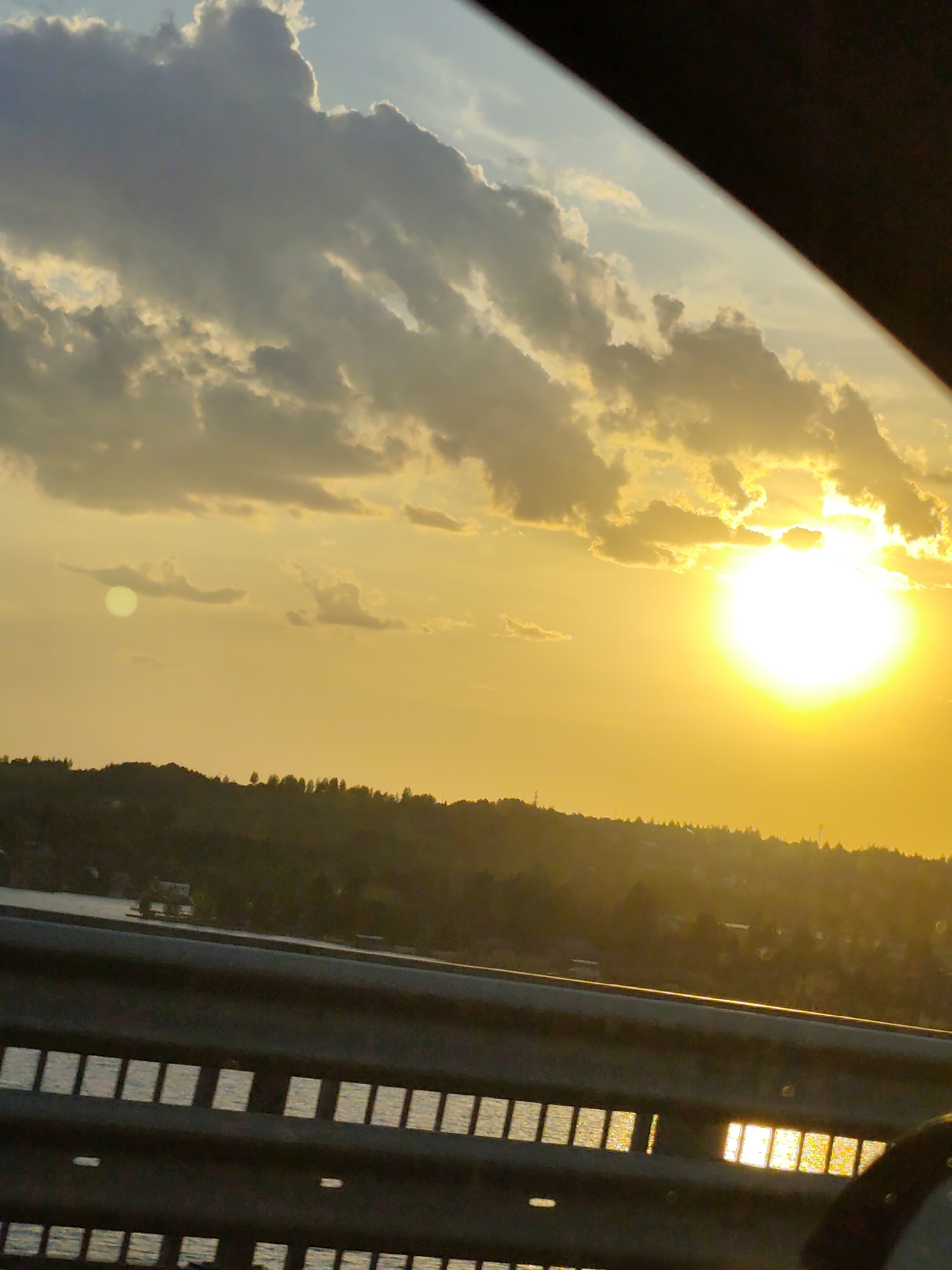 The image shows a sunset over a body of water, likely a lake or river, as viewed from a bridge. The sun is bright and partially obscured by clouds, creating a golden sky. Below the horizon, there's a tree-lined hill. A guardrail with rectangular openings lines the bottom of the frame, reflecting sunlight on the water visible through the openings. A portion of the car's interior is visible on the right and upper right of the frame. The location is identified as Пальники, Russia.