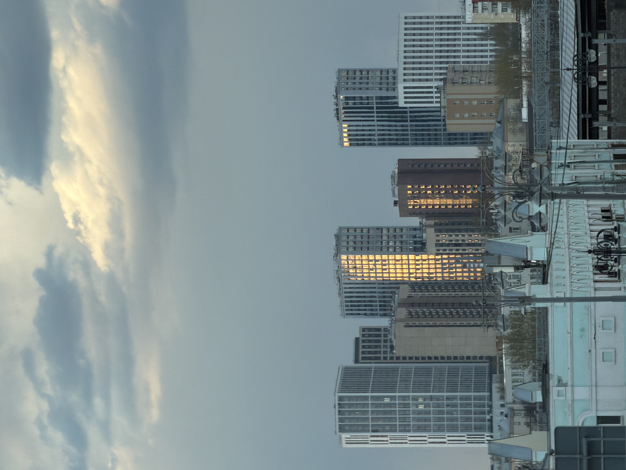 The image captures a cityscape in Moscow, Russia, under an overcast sky with striking cloud formations. The sun reflects off the windows of several tall, modern buildings, creating a golden effect. Power lines crisscross the view in the foreground. The architectural styles vary, including older, possibly Soviet-era structures, amidst contemporary designs. Street-level details are visible, including streetlights and possibly some signage.