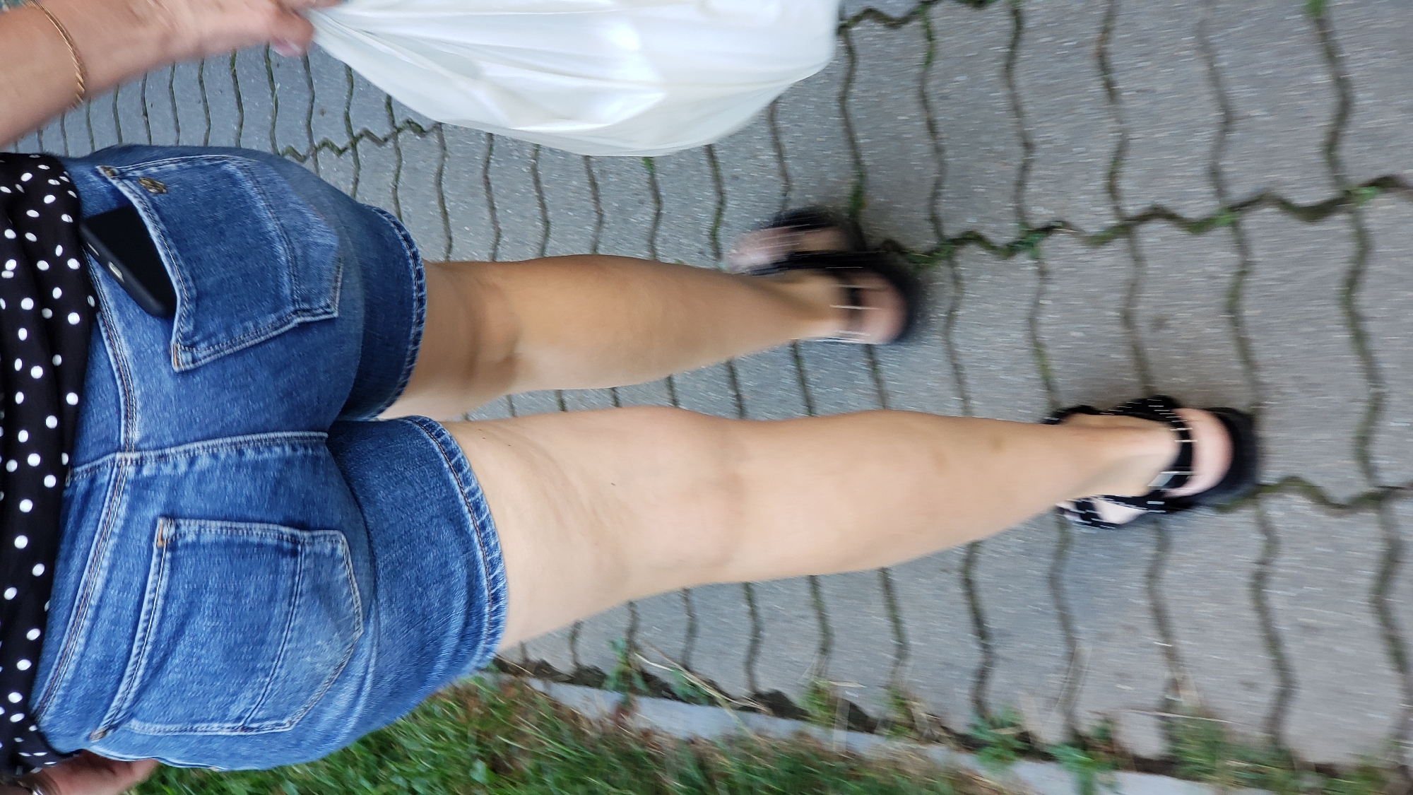 A person is walking on a paved path made of interlocking gray bricks. The path is bordered by grass and some foliage. The person is wearing denim shorts, a black and white polka dot top, and black sandals. A white bag is being carried by the person, and a smartphone is tucked into the back pocket of the shorts. The scene appears to be outdoors, possibly during the daytime, with moderate lighting conditions. The image is taken from a high-angle, slightly tilted perspective. There is no visible text in the image.