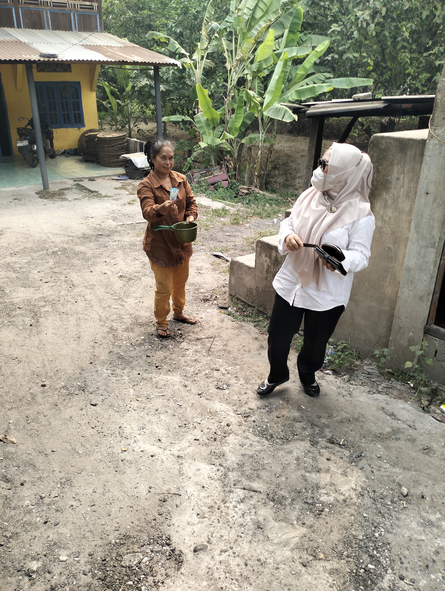 Two women are interacting in a dusty outdoor area near a simple building with a yellow facade and a corrugated metal roof. The woman on the left, wearing a brown shirt and tan pants, holds a small green cup and a few bills in her hand, appearing to be making a transaction or giving something to the other woman. She has dark hair pulled back and is looking towards the second woman. The woman on the right is wearing a white shirt, black pants, and a light-colored headscarf, along with sunglasses and a face mask. She is holding a dark-colored wallet or clutch. The environment is rural, with dirt ground, scattered stones, and lush green vegetation, including banana trees, in the background. A motorcycle is parked near the building. The scene is likely in Mandiangin, Indonesia, given the description of the location.
