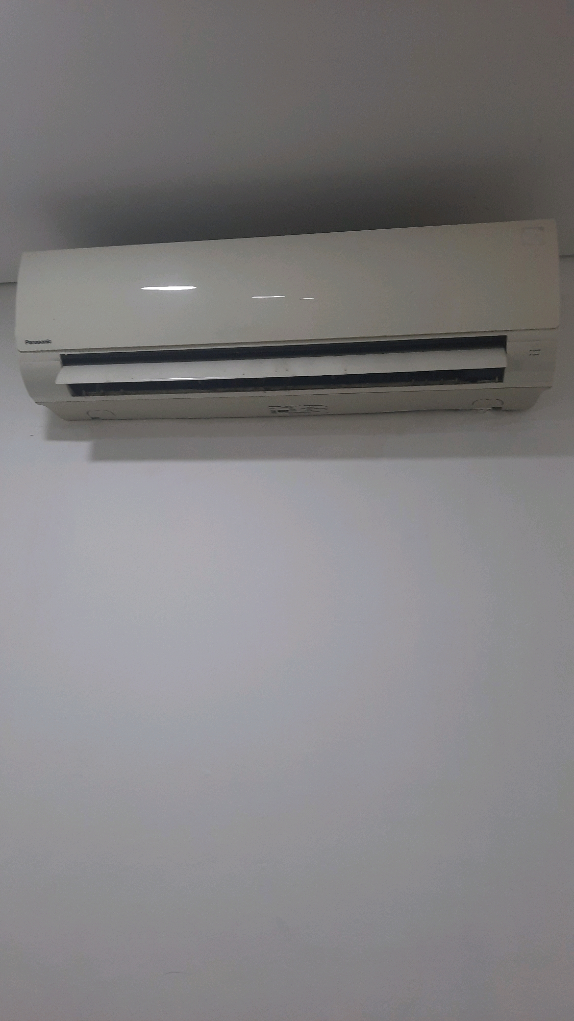 An indoor air conditioning unit is mounted high on a plain white wall, just below a grey ceiling. The unit is off-white and appears to be a split system indoor unit, with a visible vent at the bottom and a horizontal slatted vent across the front. The "Panasonic" logo is visible on the left side of the unit. The wall is a uniform, light grey, and the ceiling is a darker shade of grey, creating a minimalist and clean environment. The lighting suggests it is daytime, but the source is not directly visible. The image is static and does not depict any people or ongoing activity.