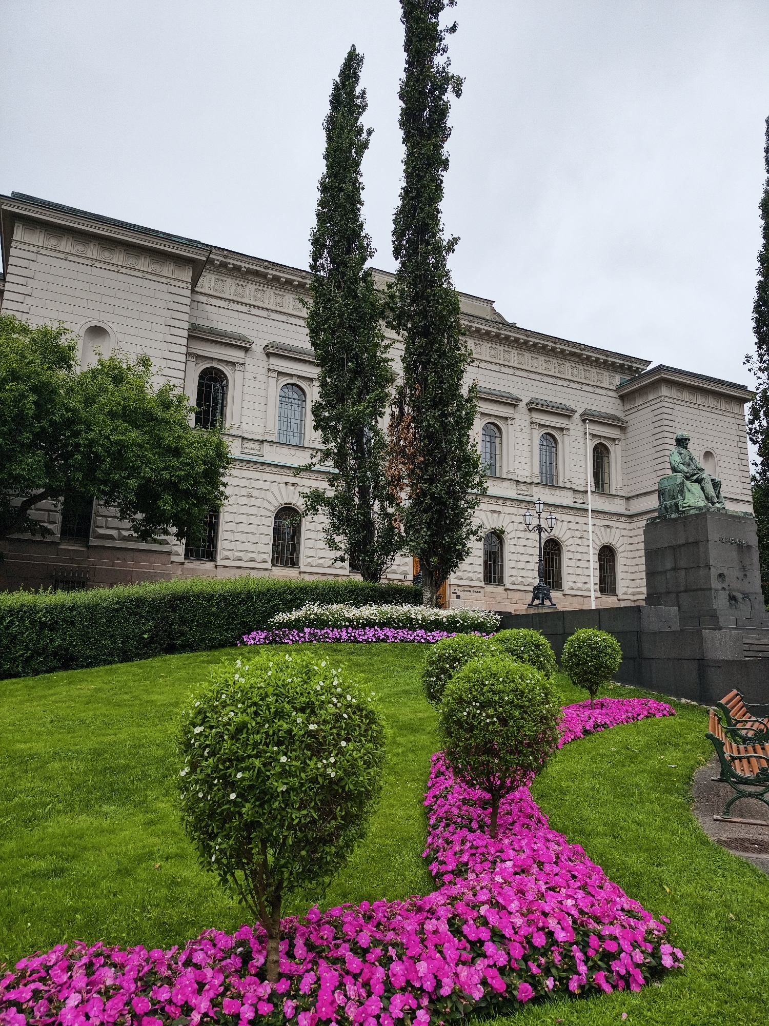 The image shows the exterior of the Finnish Parliament House in Helsinki, Finland. The building is a large, light-colored stone structure with classical architectural elements, including arched windows and decorative friezes. In front of the building is a well-maintained garden with a lush green lawn, neatly trimmed hedges, and colorful flower beds filled with bright pink and white flowers.
Several decorative spherical trees are planted in the garden, adding to the aesthetic appeal. To the right of the garden, a large bronze statue of a seated figure on a stone pedestal is visible, with the inscription "J.V. Snellman" on the pedestal. The statue is positioned in front of a dark stone wall, which appears to be part of the building's foundation or a surrounding structure.
In the foreground, a park bench with wooden slats and green metal legs is placed on the grass, inviting visitors to rest and enjoy the surroundings. The sky is overcast, suggesting a cloudy day. The overall scene depicts a serene and well-landscaped public space adjacent to an important governmental building.