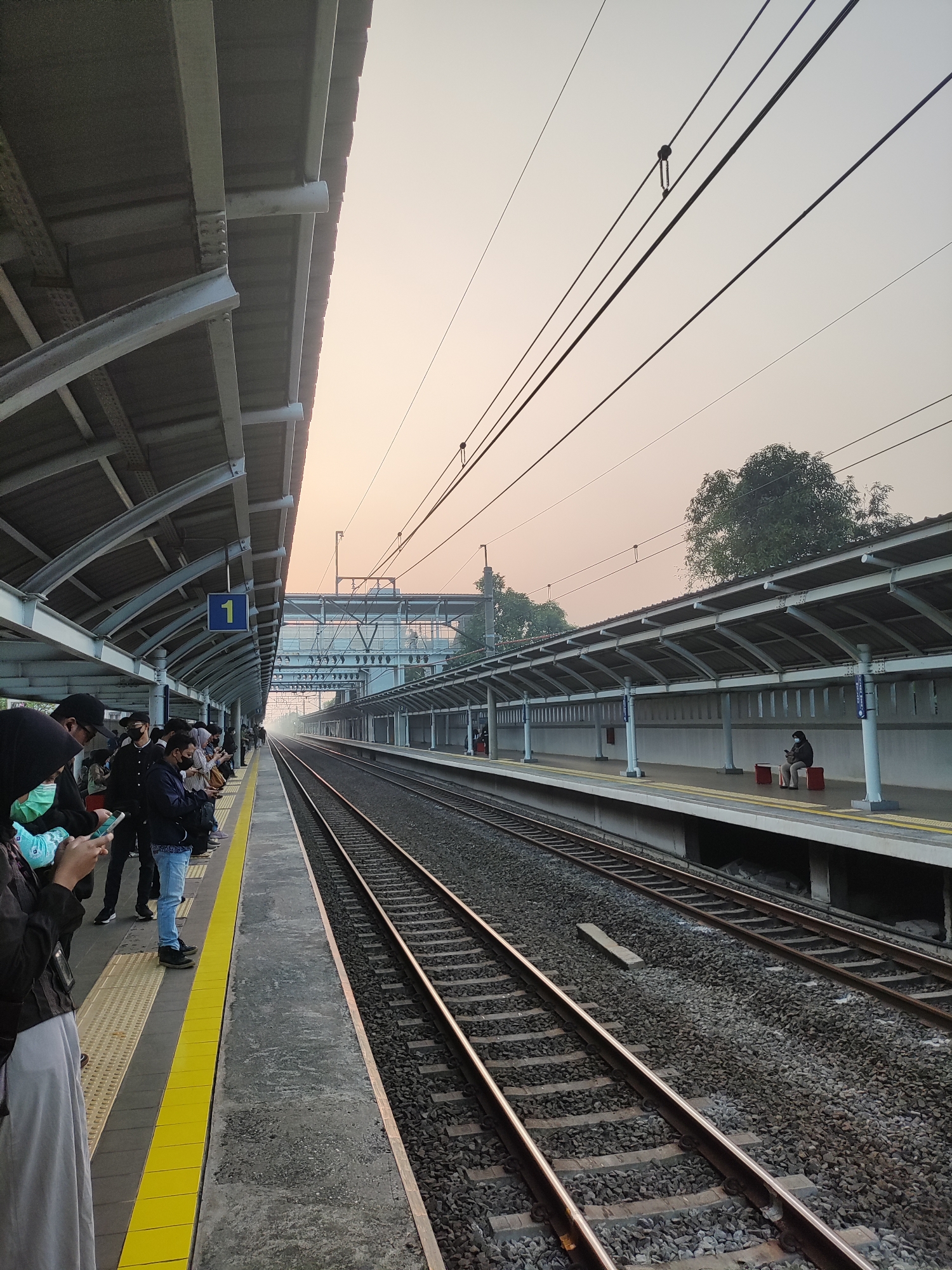The image depicts a train station platform in Telagamurni, Indonesia, during what appears to be early morning or late afternoon due to the soft, diffused light. The sky is a hazy orange-pink, and there is a visible layer of smog or dust in the air, obscuring the distant background and trees.
On the left side of the image, a covered platform is visible, supported by metal beams. Several people are standing on this platform, waiting. Some are looking at their phones, while others are conversing. Most are wearing masks, indicating a public health precaution. A blue sign with a yellow number "1" is mounted on the underside of the platform's roof.
The platform is adjacent to train tracks, with gravel ballast between the sleepers. The tracks stretch into the distance, disappearing into the hazy atmosphere. On the right side, another covered platform runs parallel to the tracks. This platform is less populated, with a single person sitting on a red bench, also wearing a mask and looking at their phone.
Overhead, a complex network of electrical wires and support structures for an overhead train system is visible against the hazy sky. The architecture of the platforms suggests a functional, modern train station. The overall scene conveys a sense of waiting and transit within an urban environment affected by atmospheric conditions.