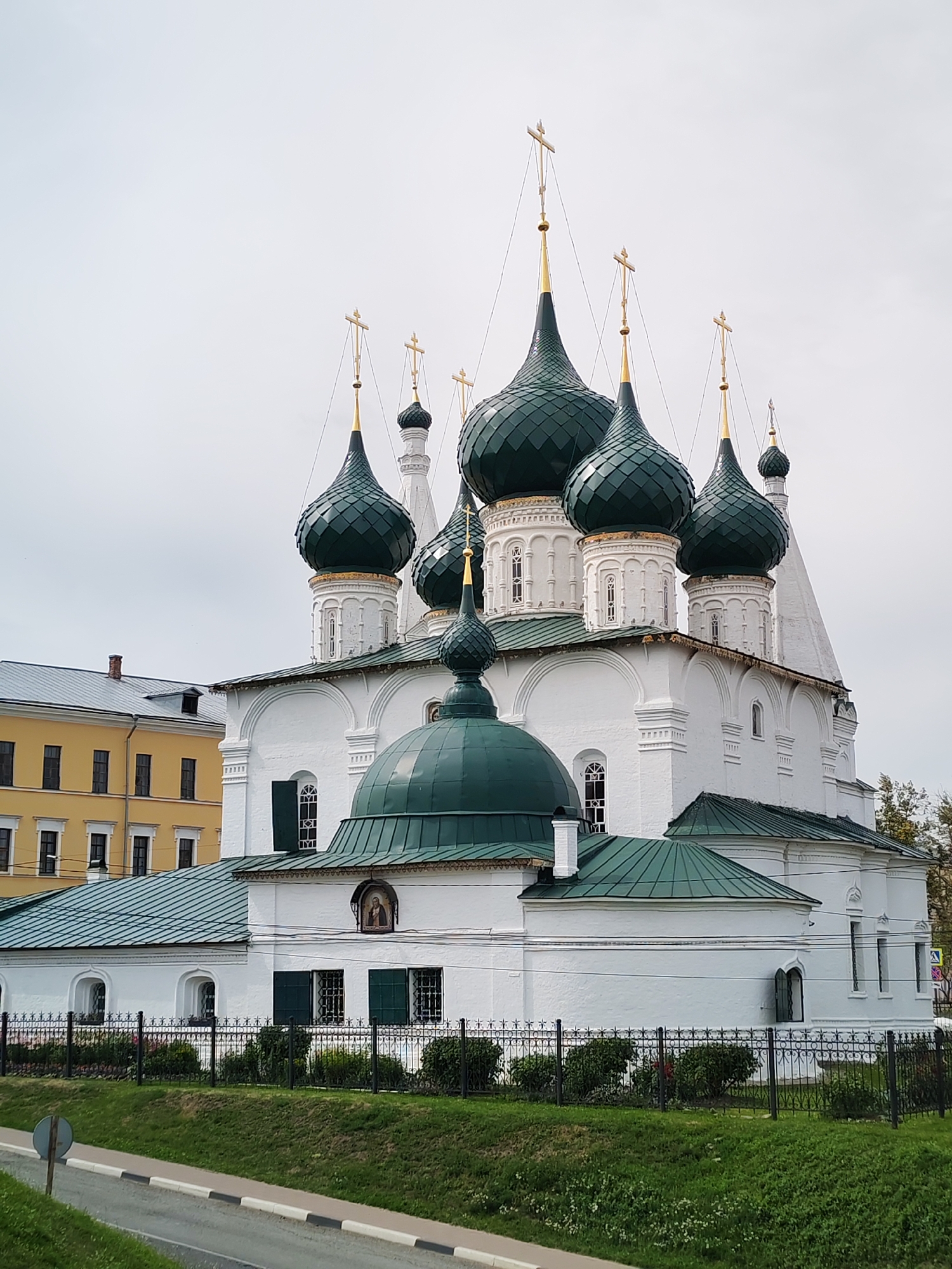 The image shows a white Orthodox church with multiple green onion domes, each topped with a golden cross. The church has several windows and ornate architectural details. In the background, a yellow building with a sloped roof is visible. The foreground features a green lawn, a black metal fence, and a paved road with a traffic sign. The sky is overcast and pale.
