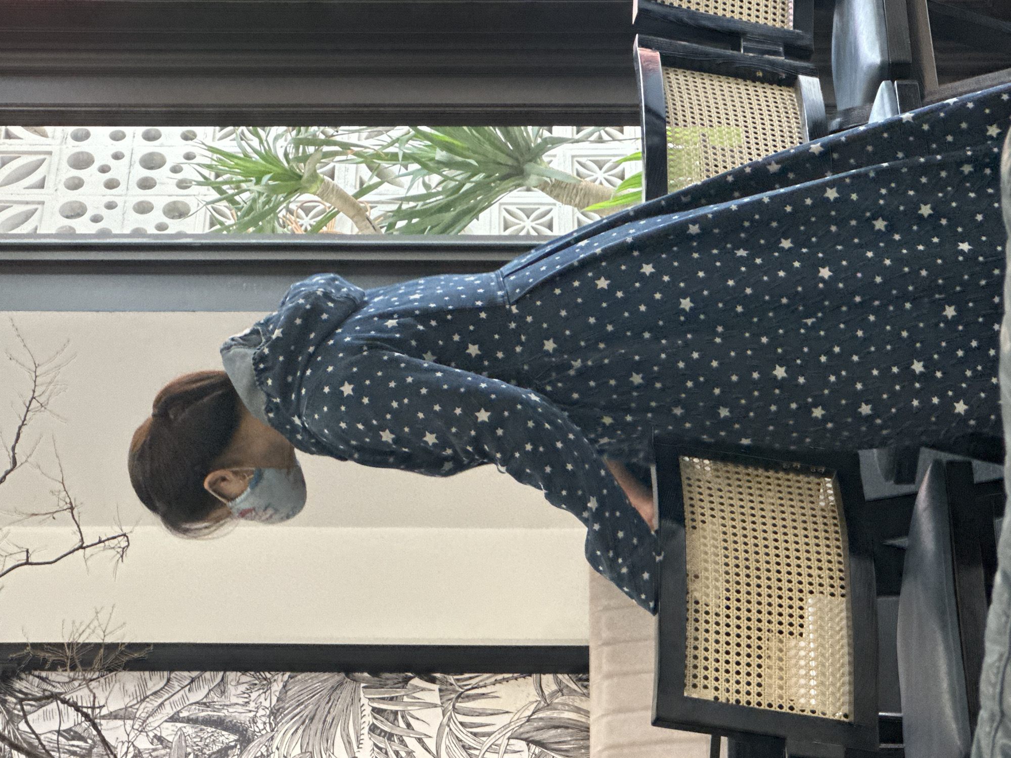 An image, rotated 90 degrees counter-clockwise, depicts a person in an indoor setting, looking downwards. The individual, likely female, has dark hair pulled back and is wearing a light blue face mask with a subtle pattern. They are dressed in a long, dark blue garment adorned with numerous small white star patterns.
The person appears to be seated or leaning forward at a table, with several dark wooden chairs featuring woven cane backs or seats visible around them and in the background. Beyond the seating area, a large window reveals an outdoor scene. Through the window, green foliage from a large plant with thick stems is visible, set against a decorative concrete block wall. This wall features a variety of geometric cutouts, including circles and stylized square/diamond patterns.
Below the window, on the interior wall, there is a large framed artwork displaying intricate black and white botanical illustrations of leaves and branches. Some sparse, dried branches are also visible in the foreground, likely part of an indoor decorative arrangement. The overall lighting suggests it is daytime, with natural light entering through the window. The architectural elements, particularly the concrete block wall, hint at a tropical or Southeast Asian location, consistent with the provided context of Phố Ràng, Vietnam. No discernible text is visible in the image.
