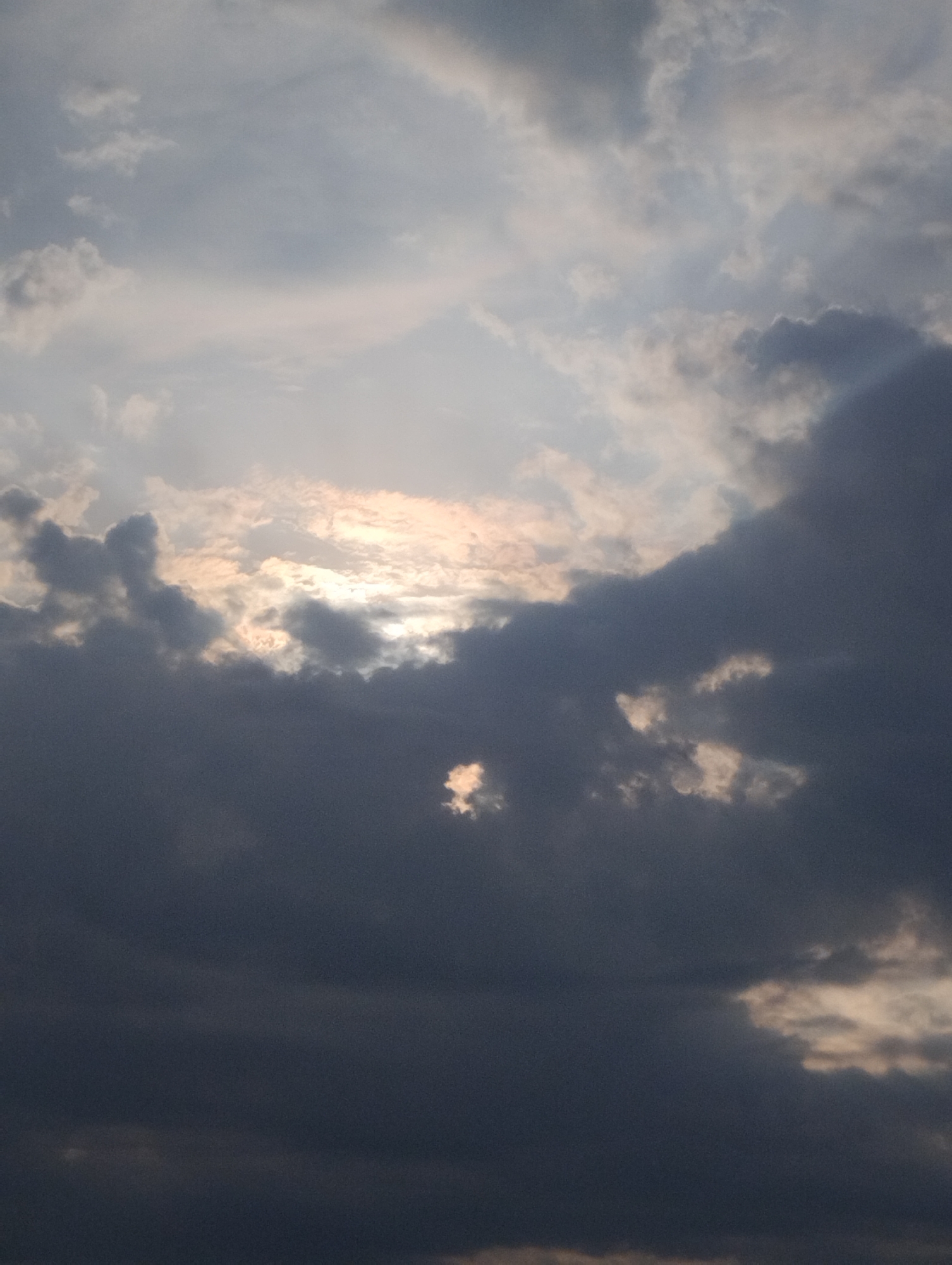 The image is a vertical shot of a cloudy sky. The clouds are dark and dramatic, with some shafts of light breaking through. The lighting suggests either sunrise or sunset. The image is not an urban landscape and does not depict any specific location in the city of Minsk, Belarus, or any other identifiable city. There are no people, subjects, objects, or actions visible in the frame. The scene is purely atmospheric.