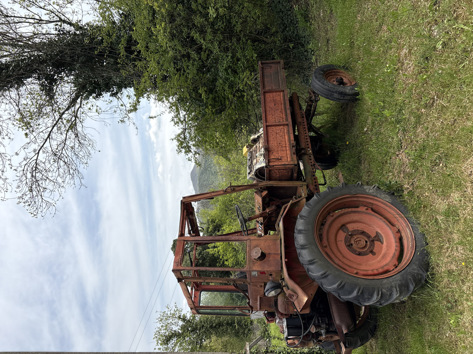 The media file displays an old, heavily rusted, and seemingly abandoned tractor situated outdoors in a rural or semi-rural setting. The tractor, predominantly a rusty red-brown color, features large rear wheels with dark tires and rusted red rims, and smaller front wheels. Its cab structure is skeletal, consisting mainly of rusted metal frames without glass, and a steering wheel is visible inside. Various engine components and mechanical parts are exposed due to the vehicle's age and decay. A rusty, box-like trailer attachment is visible behind the main body.
The tractor rests on a patch of green grass that appears somewhat overgrown. In the background, there is a dense line of trees and bushes with varied green foliage. To the left of the tractor (when oriented upright), trees with bare or sparsely leafed branches stand against the sky. In the far distance, a mountainous or hilly landscape is faintly visible beneath a bright, partially cloudy sky. A utility wire is also visible in the mid-ground.
The scene captures a static moment, with no activity or people present. The bright, diffused lighting suggests it is daytime, possibly mid-morning or afternoon, on a day with scattered clouds. The overall impression is one of neglect and the passage of time. No legible text is visible on the tractor or in the surrounding environment. This scene is located in Beregovoe, Russia.