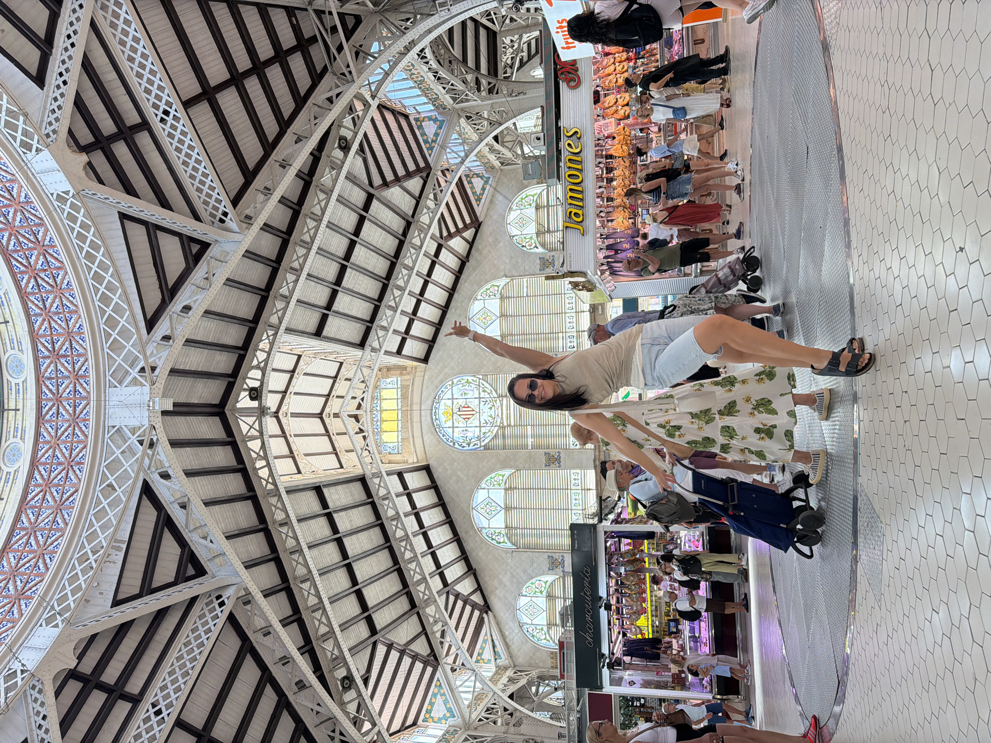 The photograph captures an interior scene of a bustling marketplace, likely a food market, beneath a grand, ornate ceiling structure. The ceiling features intricate metalwork and decorative tiling, with large arched windows allowing natural light to filter in. In the foreground, a woman in casual attire is striking a playful pose, with one arm raised and pointing upwards. She is wearing a light-colored tank top, denim shorts, and sandals. To her left, a woman in a white floral dress is pushing a blue shopping cart, and another woman in a white top and shorts stands nearby. The background is filled with stalls displaying food products, including hams hanging from the ceiling, and various shoppers browsing. The floor is tiled with a hexagonal pattern. The overall atmosphere suggests a vibrant and active public space.