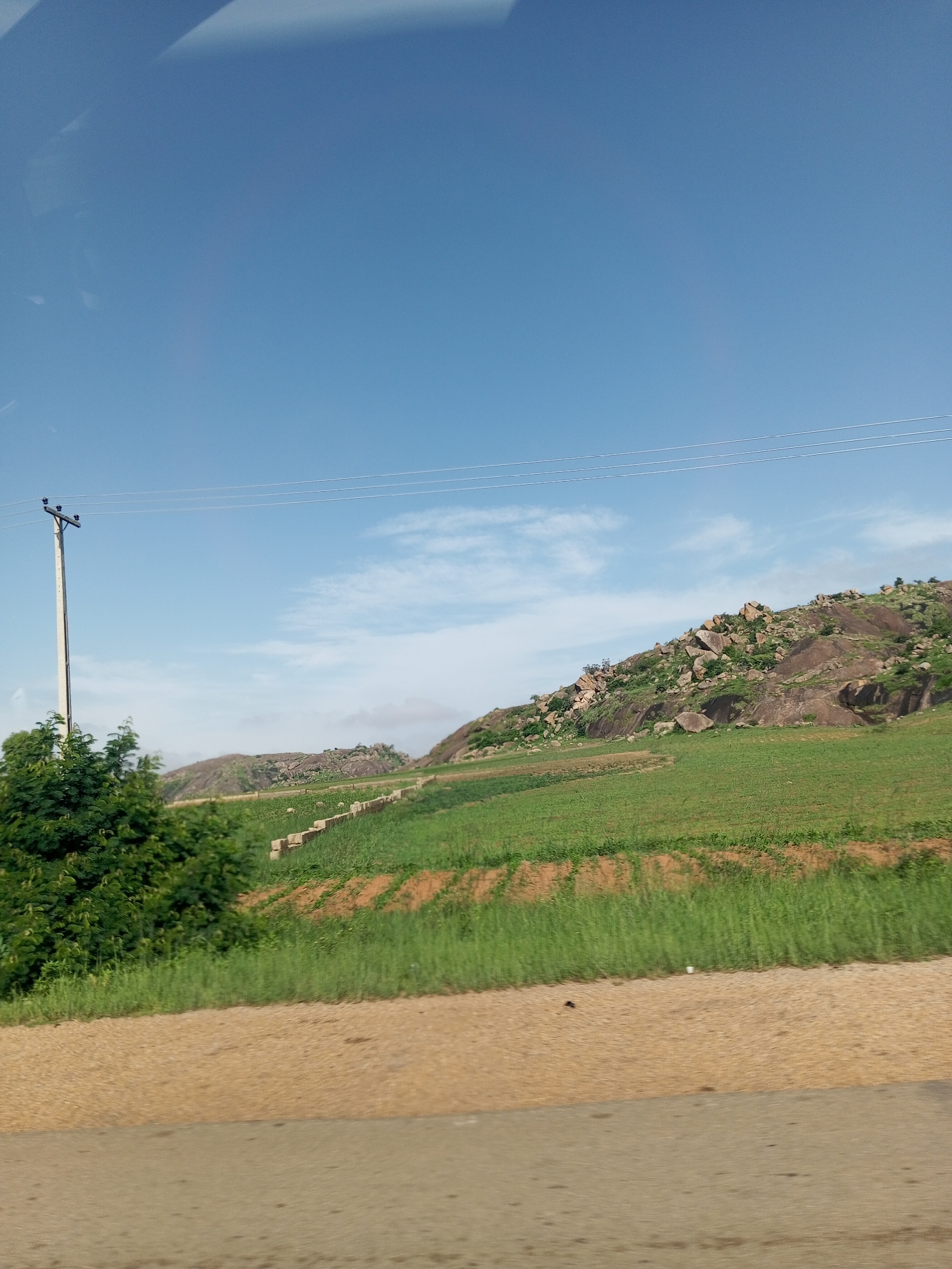 An eye-level, outdoor, medium shot captures a rural landscape under a clear blue sky in Barakin Columbai, Nigeria. The foreground features a dirt road, its surface a mix of light brown and grey tones with scattered dark debris. To the left, a dense, dark green bush occupies the edge of the frame. Beyond the bush, a tall, cream-colored utility pole stands with wires extending horizontally across the scene. The middle ground is dominated by lush green fields, with some reddish-brown soil visible in patches, suggesting agricultural activity. A low, segmented wall or terracing structure runs through the field. In the background, a rocky, sparsely vegetated hillside rises, its surface characterized by large, rounded boulders and scattered trees. The sky is a vibrant, cloudless blue, with only faint wisps of white clouds near the horizon.