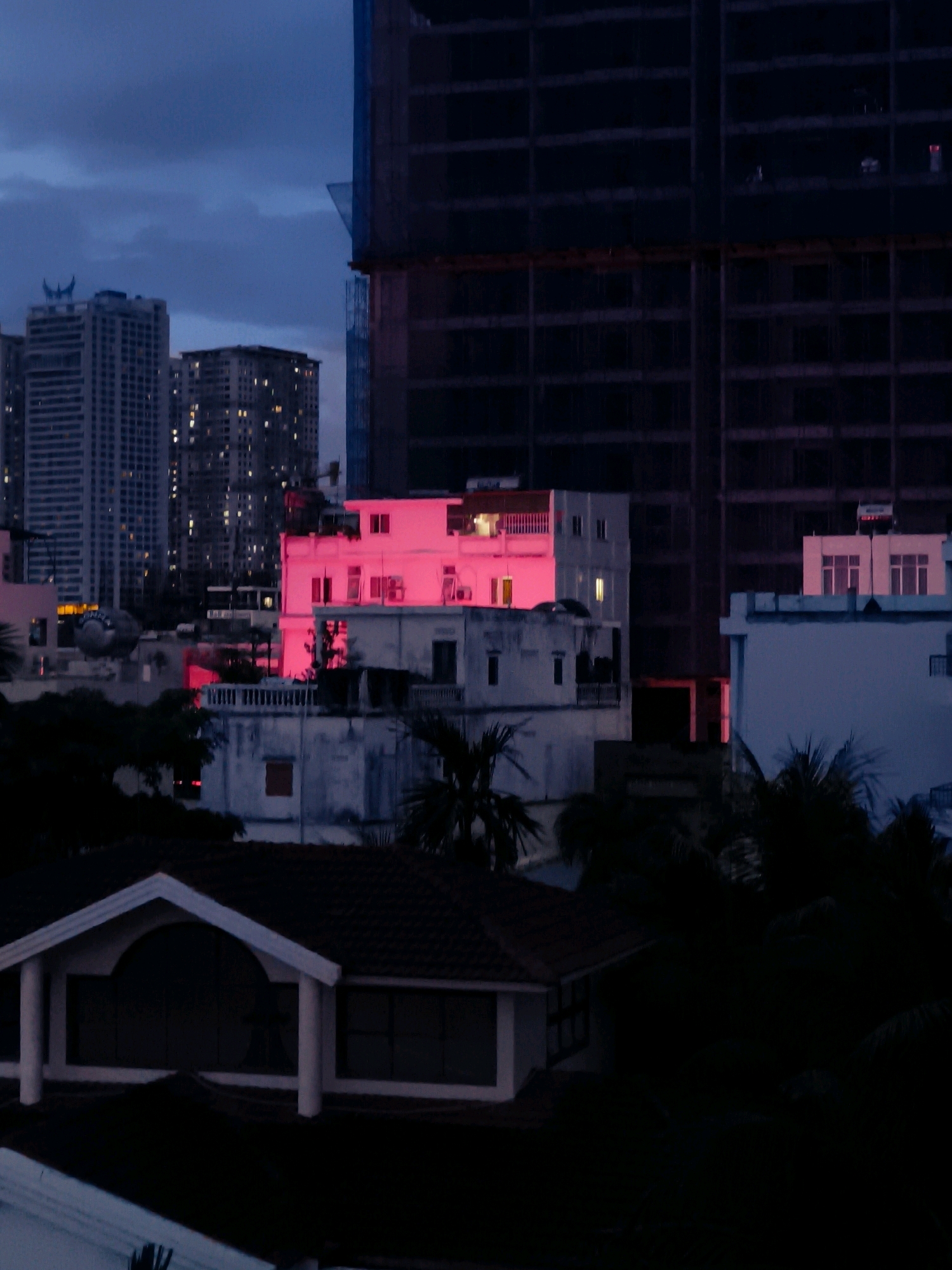 The image depicts a city skyline at dusk, with a dominant pink glow emanating from a building in the mid-ground. Several high-rise buildings are visible in the background, some with illuminated windows. A large, dark, unfinished building structure with exposed framework is situated on the right side of the image, partially obscuring the sky. In the foreground, a dimly lit residential structure with a tiled roof and a prominent white-framed entrance is visible. The overall atmosphere is twilight, with the sky transitioning from a deep blue to a darker shade. The scene is devoid of people.