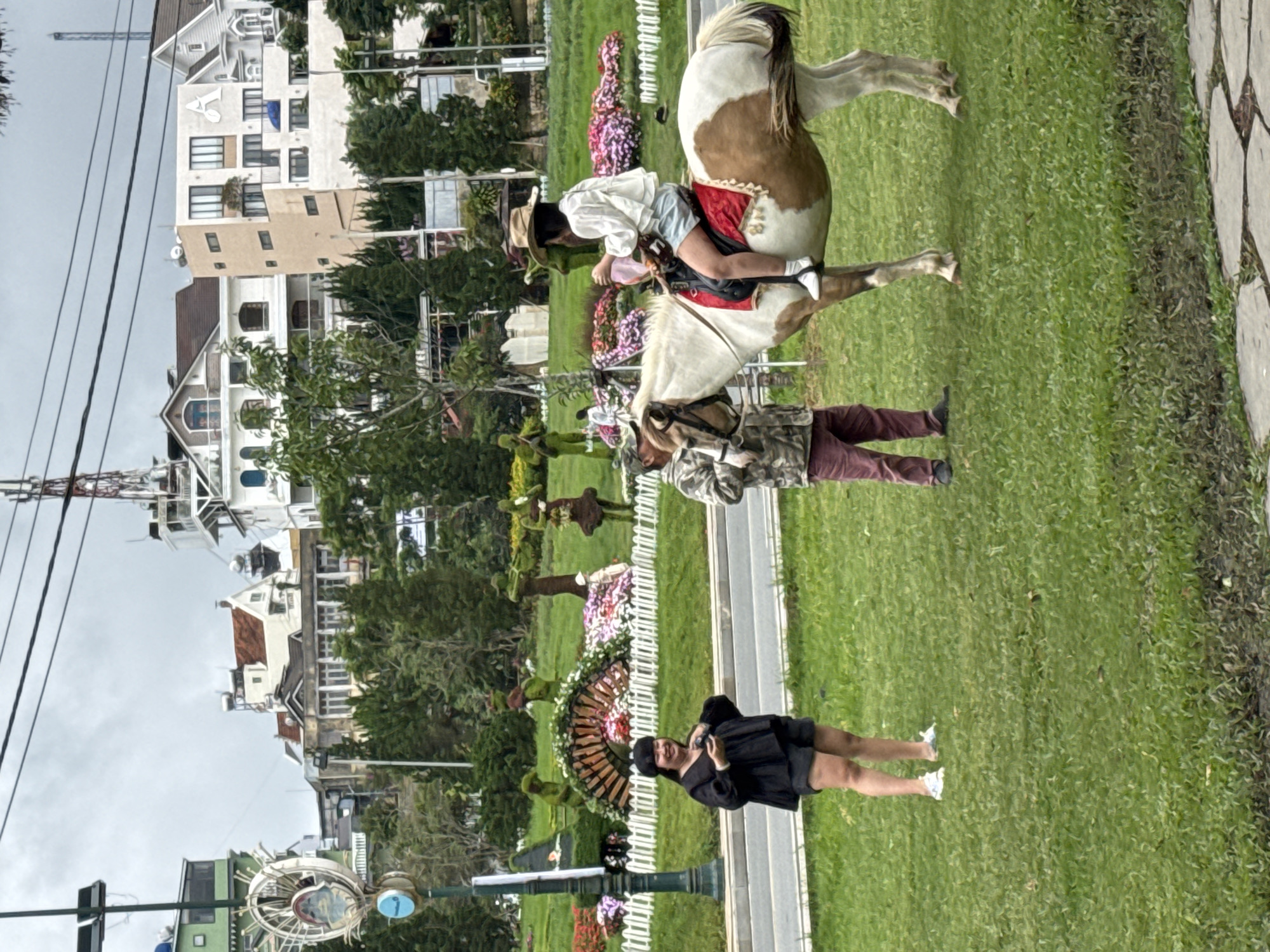 The scene depicts an outdoor park or landscaped garden in Da Lat, Vietnam, under an overcast daytime sky. Lush green grass covers most of the ground, with a paved walkway running through it.
In the mid-ground, a person wearing a cowboy hat, white long-sleeved shirt, and light shorts is seated on a brown and white horse, holding the reins. The horse is equipped with a dark saddle featuring a red decorative cloth underneath. Another individual, partially visible behind the horse, stands next to it, wearing a camouflage-patterned jacket and maroon pants. They appear to be accompanying the horse and rider.
Further down and to the left of the path, a woman with dark hair is standing, facing the camera. She is dressed in a black top, black shorts, and white sneakers, and appears to be smiling or laughing.
The park features various decorative elements, including rows of colorful flowers, some sculpted topiaries resembling animals or abstract shapes, and a large, fan-shaped floral arrangement. A white picket fence separates some of the planted areas. In the background, several multi-story buildings with varied architecture, including gabled roofs and balconies, line the horizon. On one prominent white building, a large, stylized letter, possibly "A," is visible on its facade. Utility poles and power lines are also present in the upper left of the frame. The overall impression is that of a recreational area, possibly a tourist attraction, offering horse riding and scenic views.