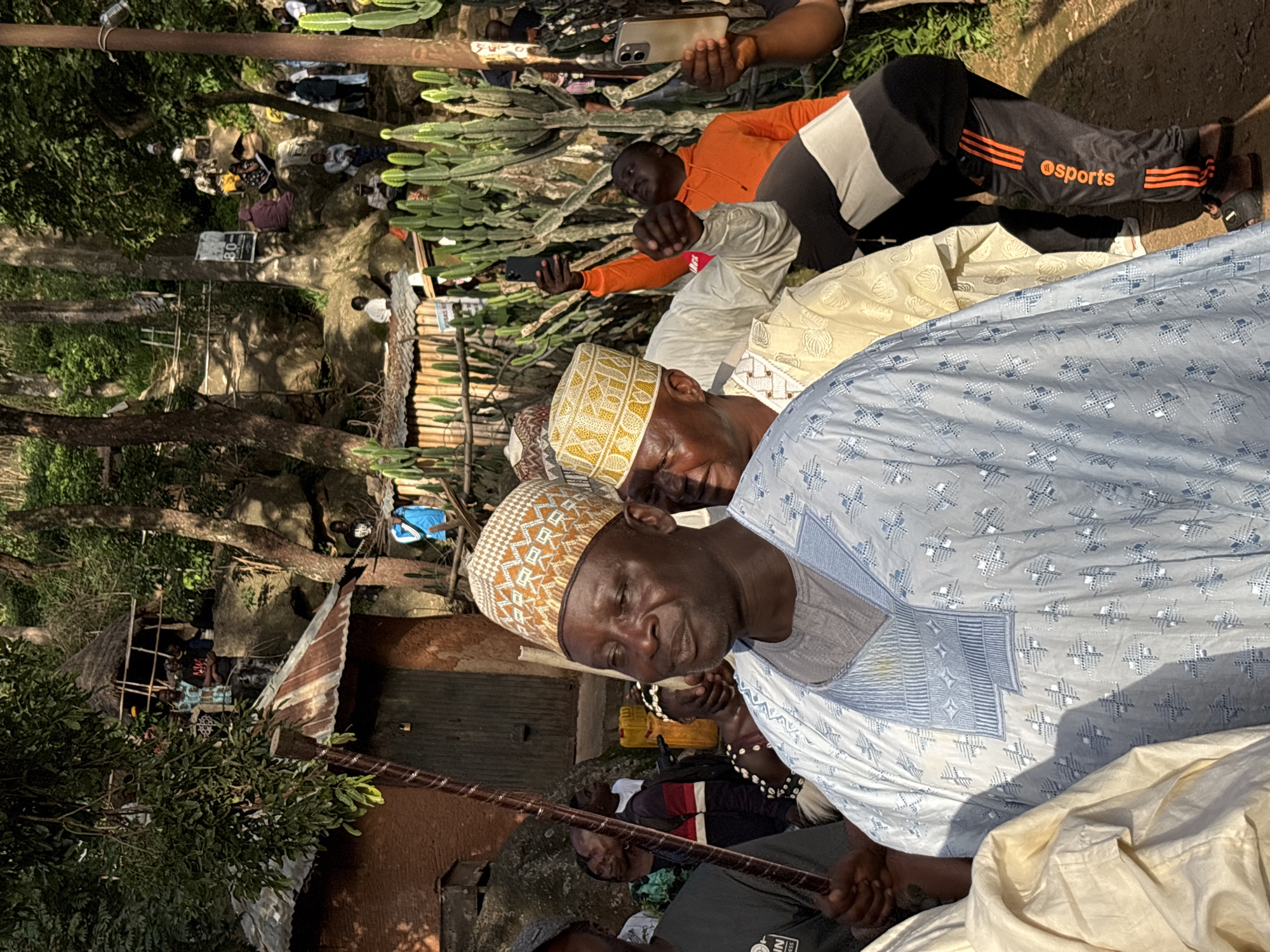 The image depicts an outdoor gathering in a hilly, wooded area that appears to be on the outskirts of Jos, Nigeria, possibly near a traditional settlement. In the foreground, a man in a light blue, embroidered tunic and a patterned cap is the central focus. He is looking slightly to his right with a pensive expression. Behind him, other individuals are visible, some wearing similar traditional attire and caps. To the right, a person in an orange hoodie and black pants with orange stripes is holding up a smartphone, likely taking a photo or video. Other people are scattered throughout the background, some seated on rocks and others standing near structures that appear to be made of corrugated metal and thatch. Lush green trees and large rocks dominate the landscape, suggesting a natural, somewhat rugged environment. The presence of several cacti-like plants also adds to the visual character of the location. The overall atmosphere seems to be that of a community event or celebration.