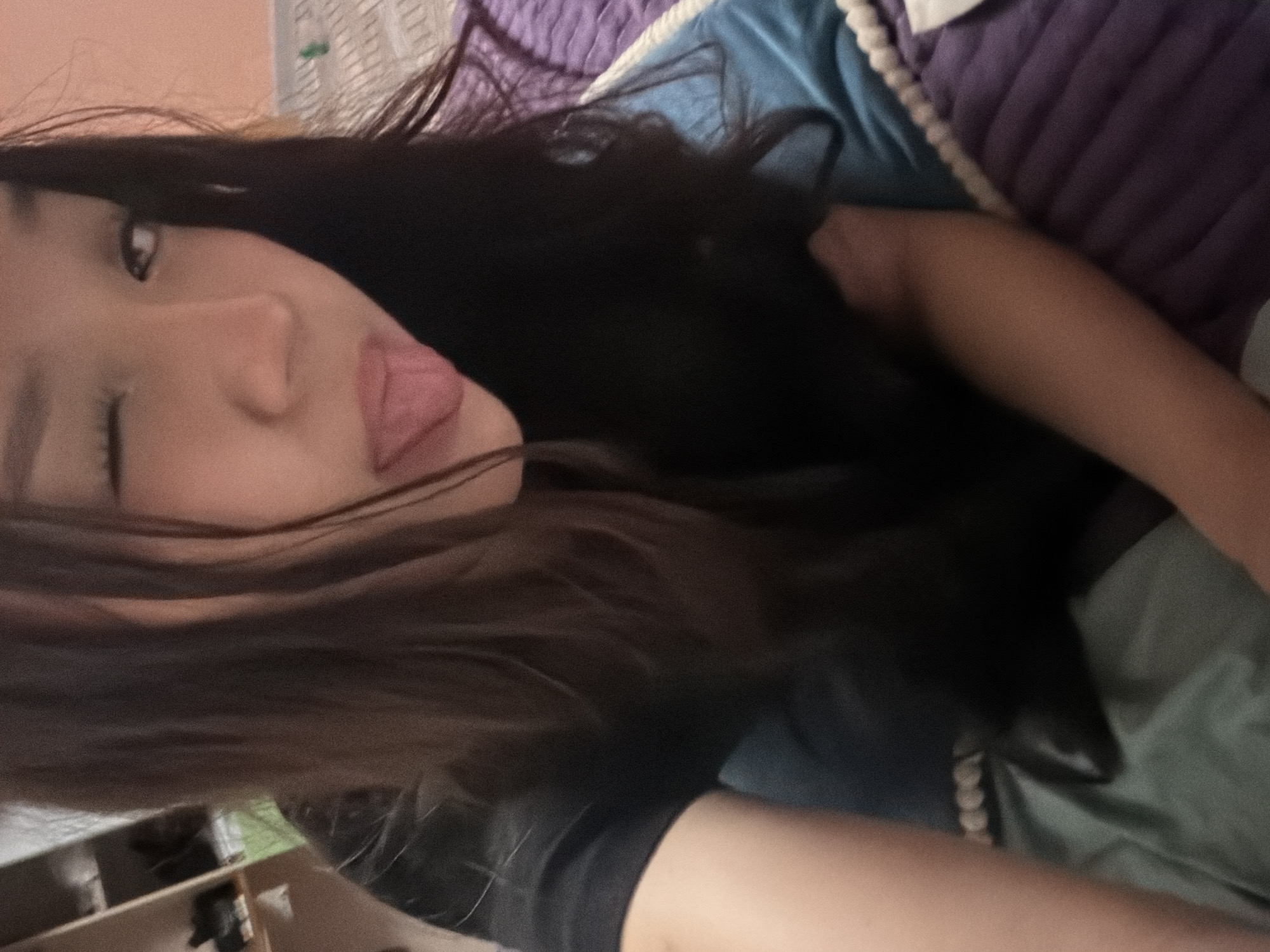 A young woman is captured in a selfie taken from a high angle, looking directly at the camera. She has long, dark brown hair that partially obscures her face. Her right eye is closed in a wink, and her tongue is sticking out. She is wearing a black t-shirt. The background shows a room with furniture, including shelves with various items on the left and a bed with a purple and blue patterned comforter. The lighting appears to be artificial and dim, creating a somewhat grainy image quality. The location is unspecified.
