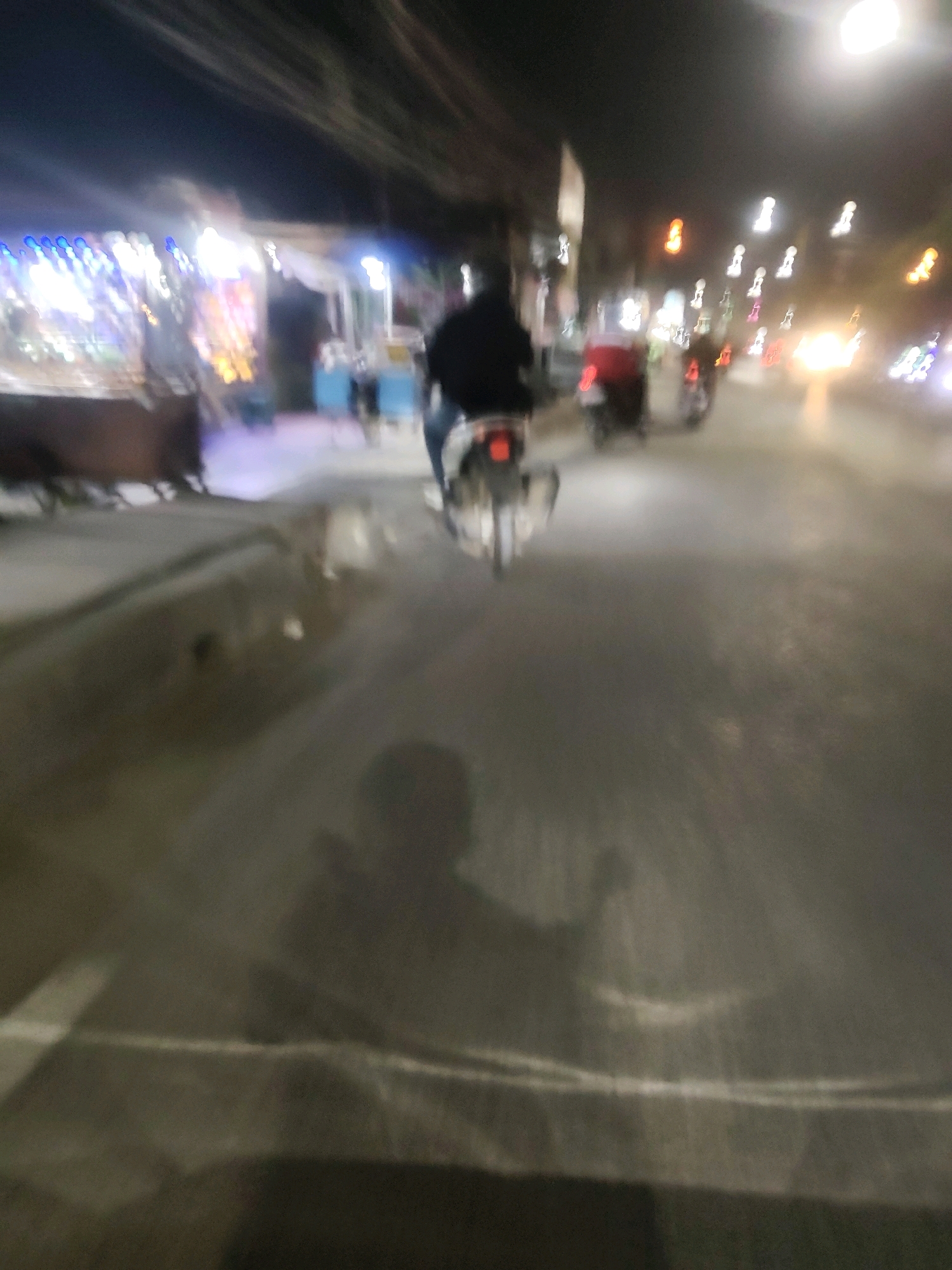The image captures a blurry nighttime scene on a city street, likely in Mekarmukti, Indonesia, as indicated by the presence of multiple motorcycles and the general urban lighting. In the foreground, a person is riding a motorcycle, their silhouette visible against the streetlights. The road surface appears wet, reflecting the lights and creating streaks of illumination. On the left side of the image, stalls with bright, colorful lights suggest a marketplace or street vendors. Further down the road, other motorcycles and vehicles are visible, some with illuminated headlights and taillights. The overall impression is one of a bustling, active street scene at night, characterized by motion blur and the vibrant glow of artificial lights.