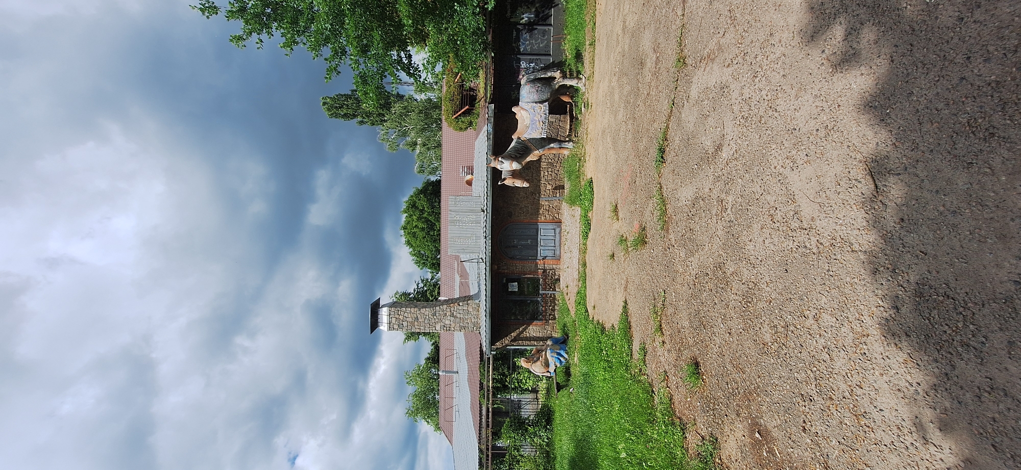 A view from a gravelly yard shows a rustic, single-story building with a stone chimney and a metal roof. Two horse statues stand near the building's entrance, one of which is saddled. A statue of a person is seated on a bench in the grassy area to the left of the building. Trees and a cloudy sky are visible in the background. The image appears to be taken in Bila Tserkva, Ukraine, though there is no explicit indication of this in the image itself.