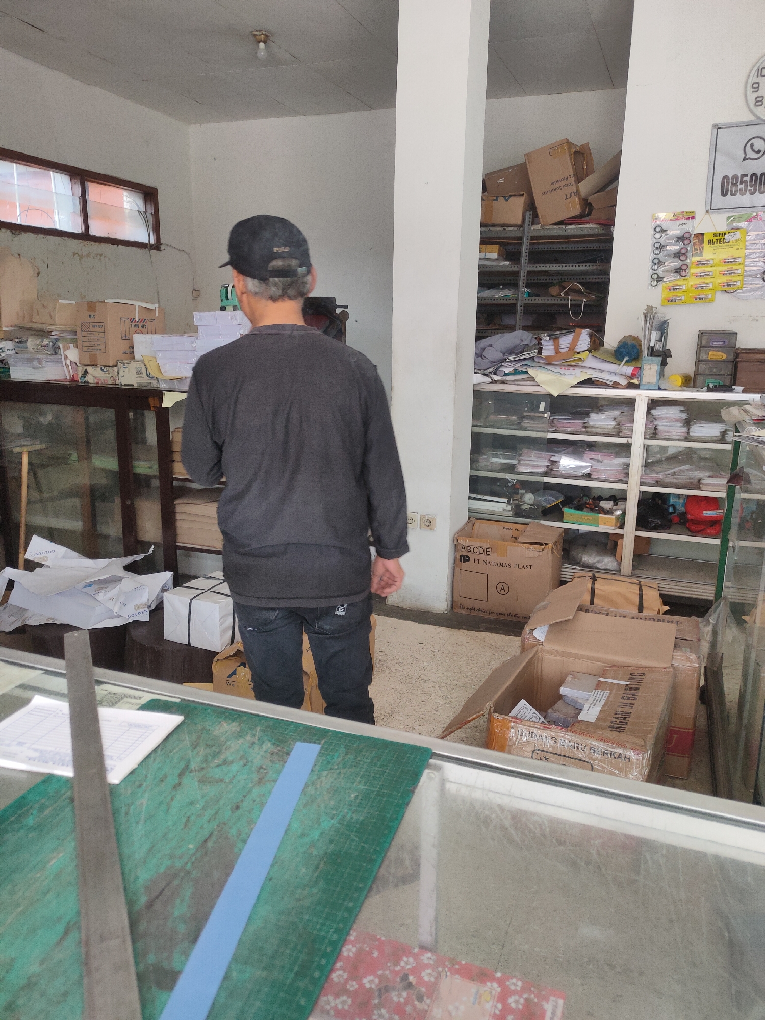 The image captures the interior of what appears to be a print shop or stationery store in Sumedang, Indonesia. A man wearing a dark baseball cap and a dark long-sleeved shirt is standing with his back to the camera, looking towards shelves stacked with paper and boxes. The store is filled with various items, including boxes of paper, office supplies, and what seem to be printing materials. In the foreground, a cutting mat with a ruler and a blue strip is visible, suggesting recent activity or preparation for work. The environment appears somewhat cluttered but functional, typical of a small business establishment.