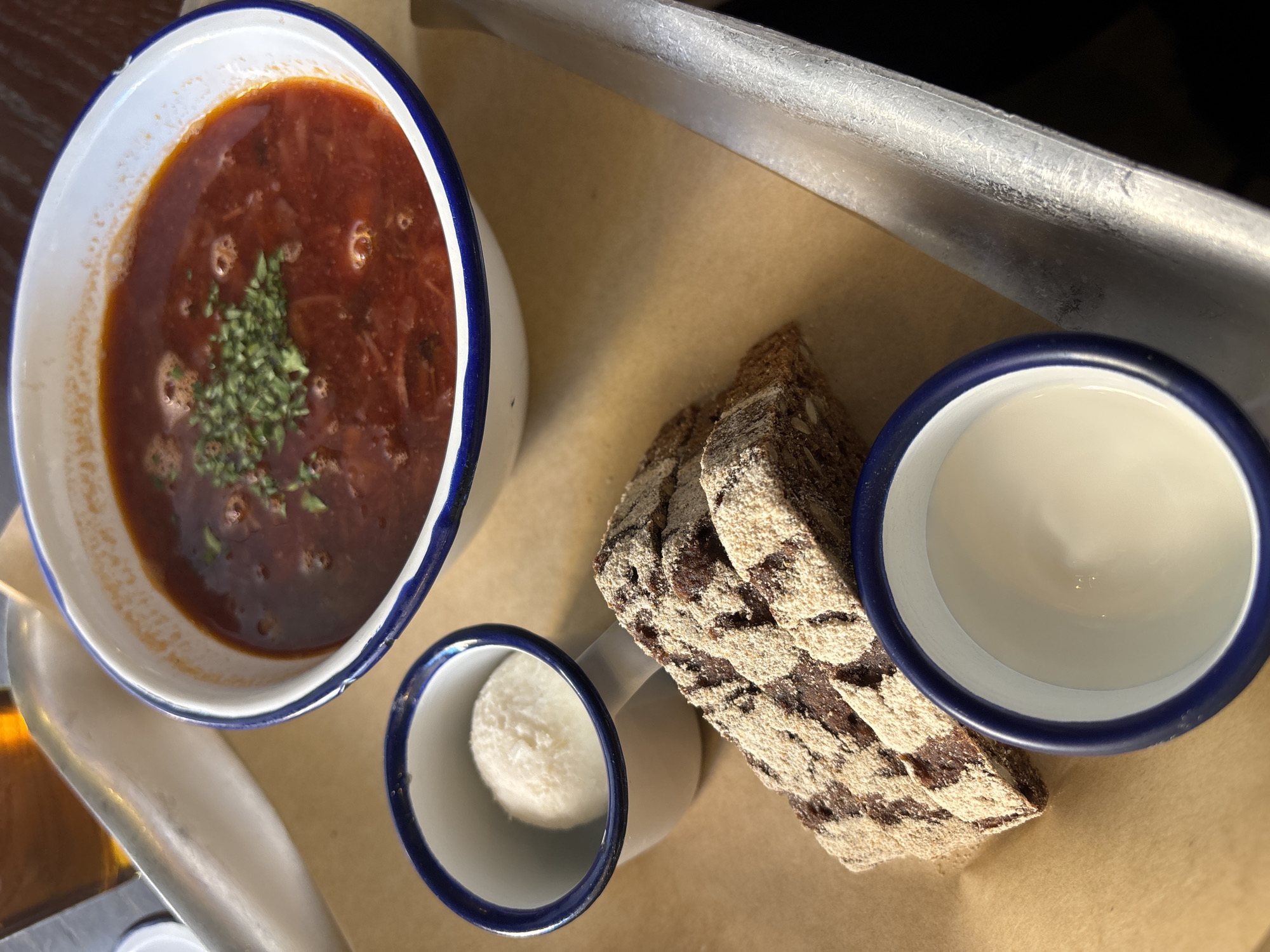 The image displays a meal served on a rectangular metallic tray, which is lined with light brown craft paper. The primary subject is a white enamelware bowl with a distinct blue rim, filled with a deep red borscht soup. The soup's surface features small bubbles and is garnished with a sprinkle of finely chopped green herbs, likely dill or parsley.
Accompanying the soup are several items. To the lower left, a smaller white enamelware mug, also with a blue rim and a handle, contains a spherical scoop of white, thick, creamy substance, identifiable as smetana (sour cream). Adjacent to and partially beneath the soup bowl are two slices of dark brown rye bread, characterized by a lighter, patterned crust and visible seeds. Below these bread slices, a third small white enamelware cup with a blue rim holds a shallow quantity of a light-colored liquid, likely kvass.
In the blurred background, towards the upper left, an out-of-focus object appears to be a glass or bottle containing an amber liquid. The overall presentation, including the specific food items (borscht, smetana, dark rye bread) and the use of enamelware and craft paper, is characteristic of the "Stolovaya №1 Kopeyka" (Столовая №1 Копейка) chain of budget-friendly canteens or cafes found across Saint Petersburg, Russia. The scene depicts a prepared meal ready for consumption in a casual dining environment within such an establishment.