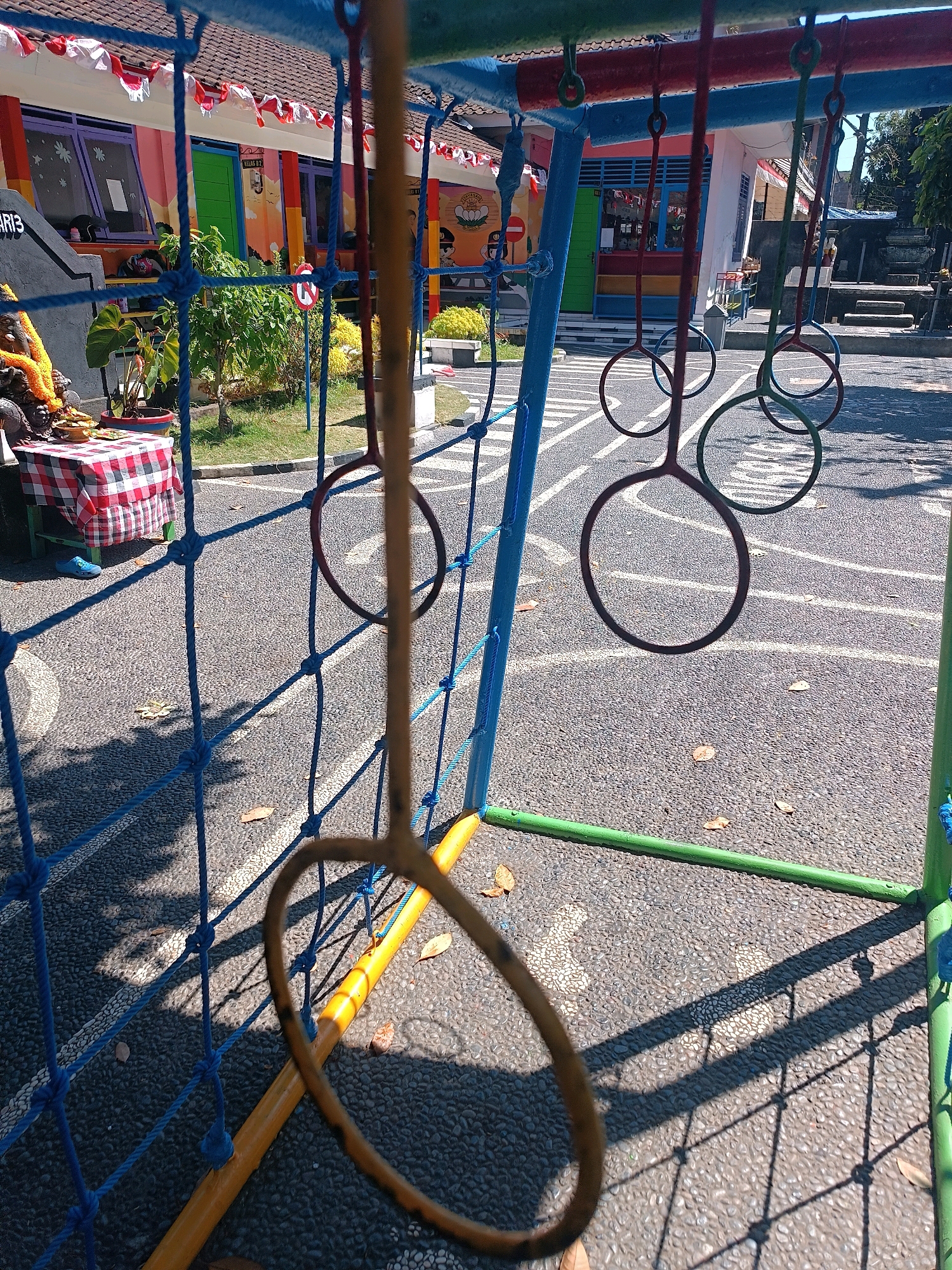The image depicts an outdoor playground scene, likely at a school in Tabanan, Indonesia. A blue net, part of a climbing structure, dominates the foreground. Hanging from a metal bar above are several rings, some dark red and others green, suspended by thin cords. One ring, a tan or light brown color, is prominently in focus in the center of the frame, casting a distinct shadow on the ground. The ground itself is asphalt with white markings, possibly a track or play area. In the background, a colorful building with purple-framed windows and a green door is visible. Decorative red and white banners are strung along the building's facade. To the left, a small shrine with a statue adorned with orange flowers sits on a table covered with a red and white checkered cloth. A single blue shoe is on the ground near the table. The environment appears sunny, with shadows from the playground equipment and net clearly visible on the asphalt.