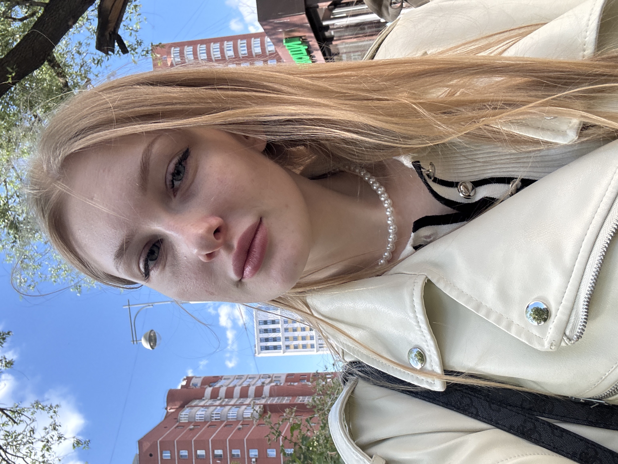 A young Caucasian woman with long blonde hair and blue eyes, wearing a white leather jacket and a pearl necklace, takes a selfie outdoors on a sunny day. She is standing in front of tall, modern buildings, suggesting an urban environment. The buildings appear to be apartment complexes, with one being predominantly red and white, and another lighter colored with many windows. Trees with green foliage are visible, with branches extending into the frame, and a streetlamp is positioned in the background. The sky is blue with scattered white clouds. The overall scene suggests a casual day out in a city that could be Perm, Russia, given the architectural style of the buildings.