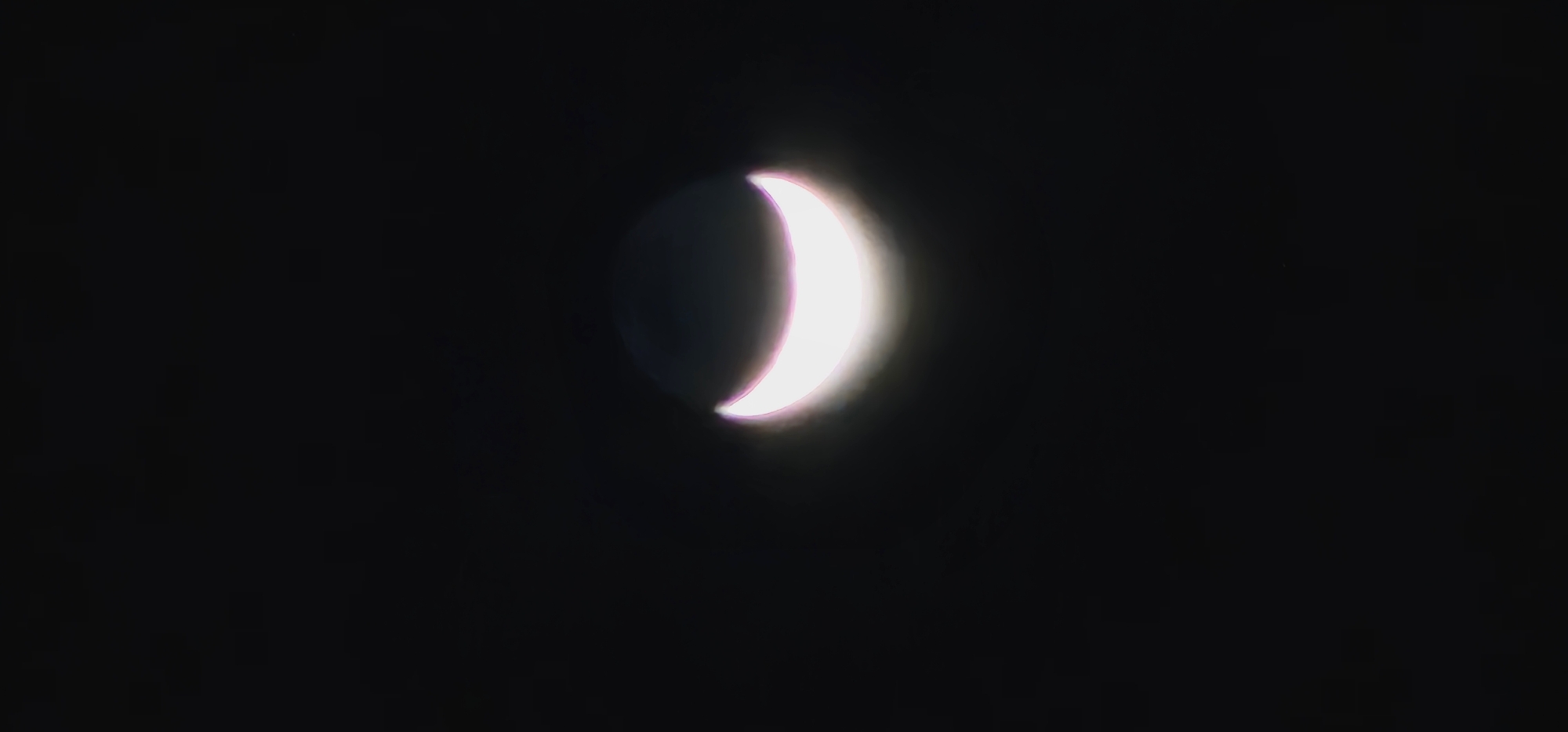 A crescent moon shines brightly against a black sky. The moon is a thin sliver of light, with a halo surrounding it. The location is not discernible from the image provided. No people, subjects, objects, actions, interactions, or specific environmental features of Denpasar, Indonesia, are visible.