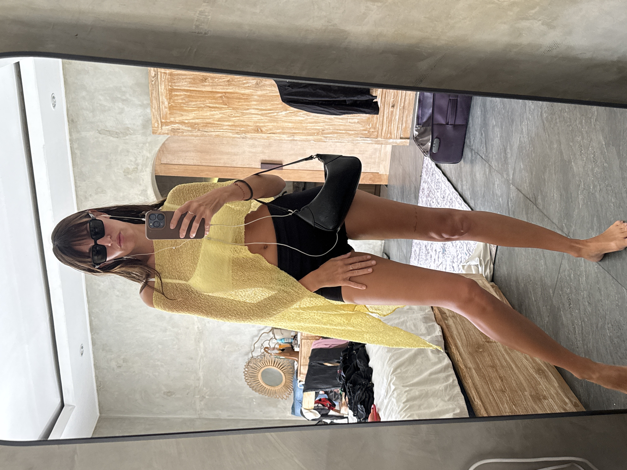 A full-length, medium shot captures a young woman in a bedroom, taking a mirror selfie. She is positioned in the center of the frame, facing the mirror, her body angled slightly to the left. She is wearing a bright yellow, sheer, off-the-shoulder top over a yellow bra and black shorts. Her arms are extended, holding a smartphone up to take the photo. White earbuds are dangling from her ears. She has dark brown hair with bangs and is wearing black sunglasses. A small black handbag is slung over her left shoulder.
The background of the reflection reveals a bedroom. To the left, a bed with white linens is visible, partially covered with folded clothes. A wooden dresser with a round, woven mirror hangs above it, adorned with decorative branches and bottles. To the right, a large wooden wardrobe stands open, revealing some clothing inside. The floor is tiled with dark gray tiles, and a patterned rug lies near the bed. A purple suitcase is open on the floor in the background. The overall aesthetic suggests a bohemian or tropical style. The scene appears to be in a private residence, possibly a hotel room or guesthouse, in North Kuta, Indonesia.