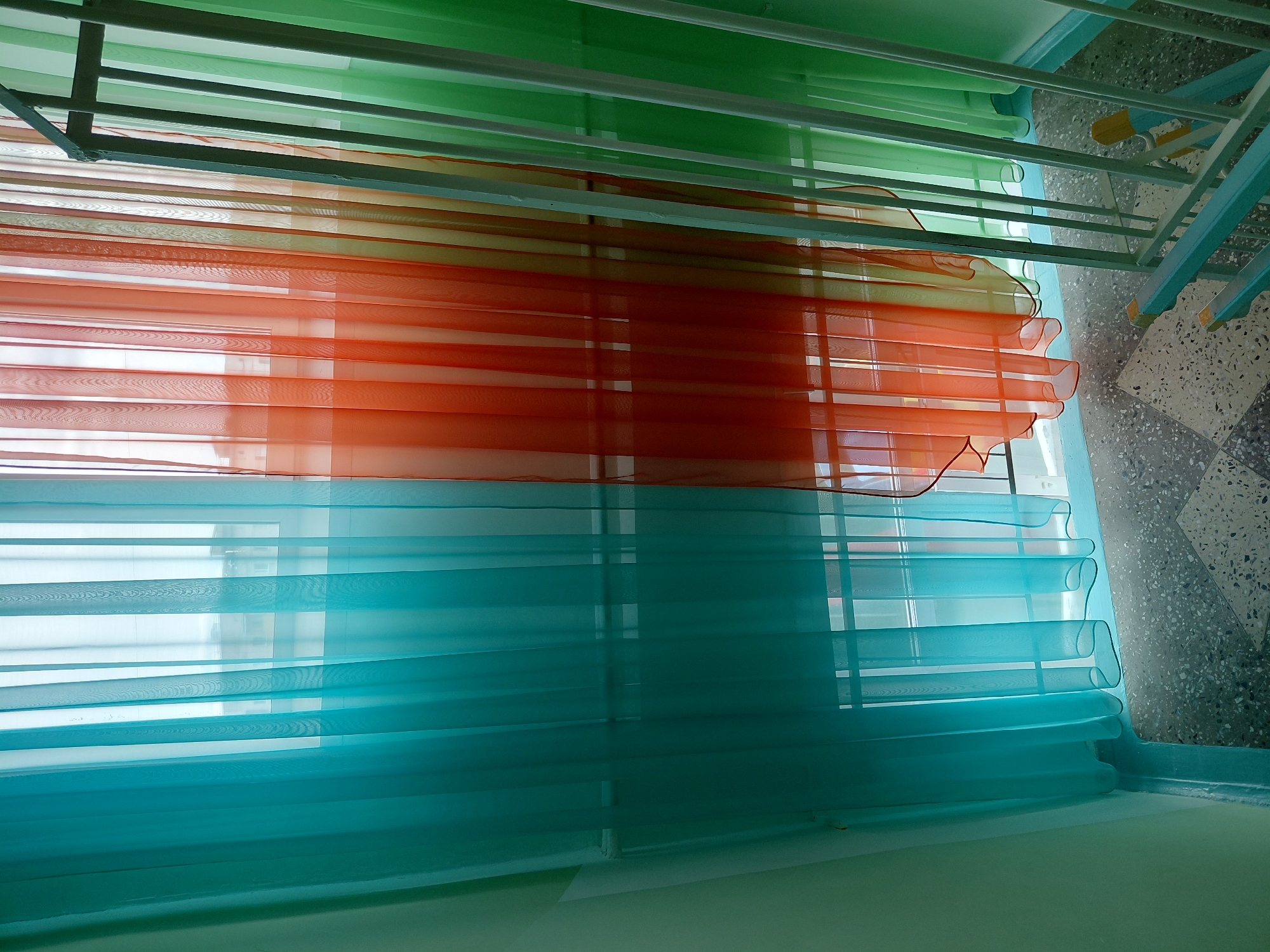 The image displays a vibrant, multi-colored set of translucent blinds, likely seen from inside a room looking outwards. The blinds are arranged horizontally in three distinct color sections. At the top, there is a band of bright green sheer fabric. Below this, a wide section of orange or red sheer fabric is visible, characterized by wavy, ruffled edges in some parts. The lowest section features a striking teal or blue sheer fabric, also exhibiting similar ruffled or folded edges.
Through the translucent material of the blinds, faint, blurred shapes and lines suggest a window or another interior space beyond, allowing light to filter through. To the right of the blinds, parts of a structure are visible. This includes white metal or plastic bars, possibly part of a drying rack, window frame, or balcony railing. Below this structure, a speckled gray and white tiled floor can be seen, which further suggests a balcony, sunroom, or a hallway with a large window.
No people are present in the image, and no specific activity is taking place; the scene is static. The bright illumination implies it is daytime. The image appears to be taken from a slightly low angle, looking up towards the top of the blinds and the ceiling area. There is no visible text in the image.