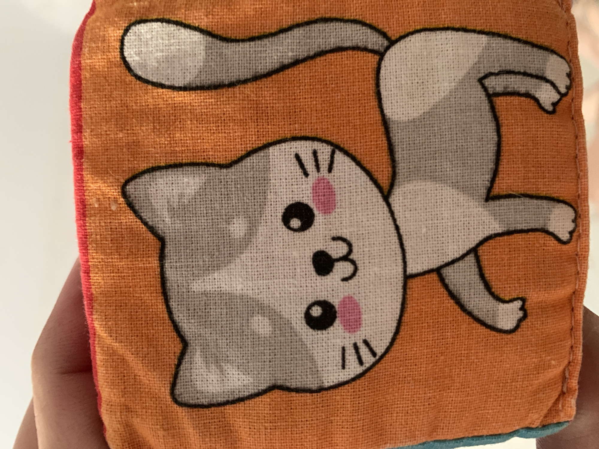 A hand is holding a fabric cube with a cartoon cat printed on it. The cat is gray and white with black outlines. It has large, round eyes, a small nose, a smiling mouth, and pink blush marks on its cheeks. The cat's body is depicted in a playful pose, with its tail arched upwards. The background of the cube is a vibrant orange fabric. The cube is a soft toy, likely for a baby or child. The scene is indoors, with a white background visible behind the cube and hand. There are no visible text elements in the image.