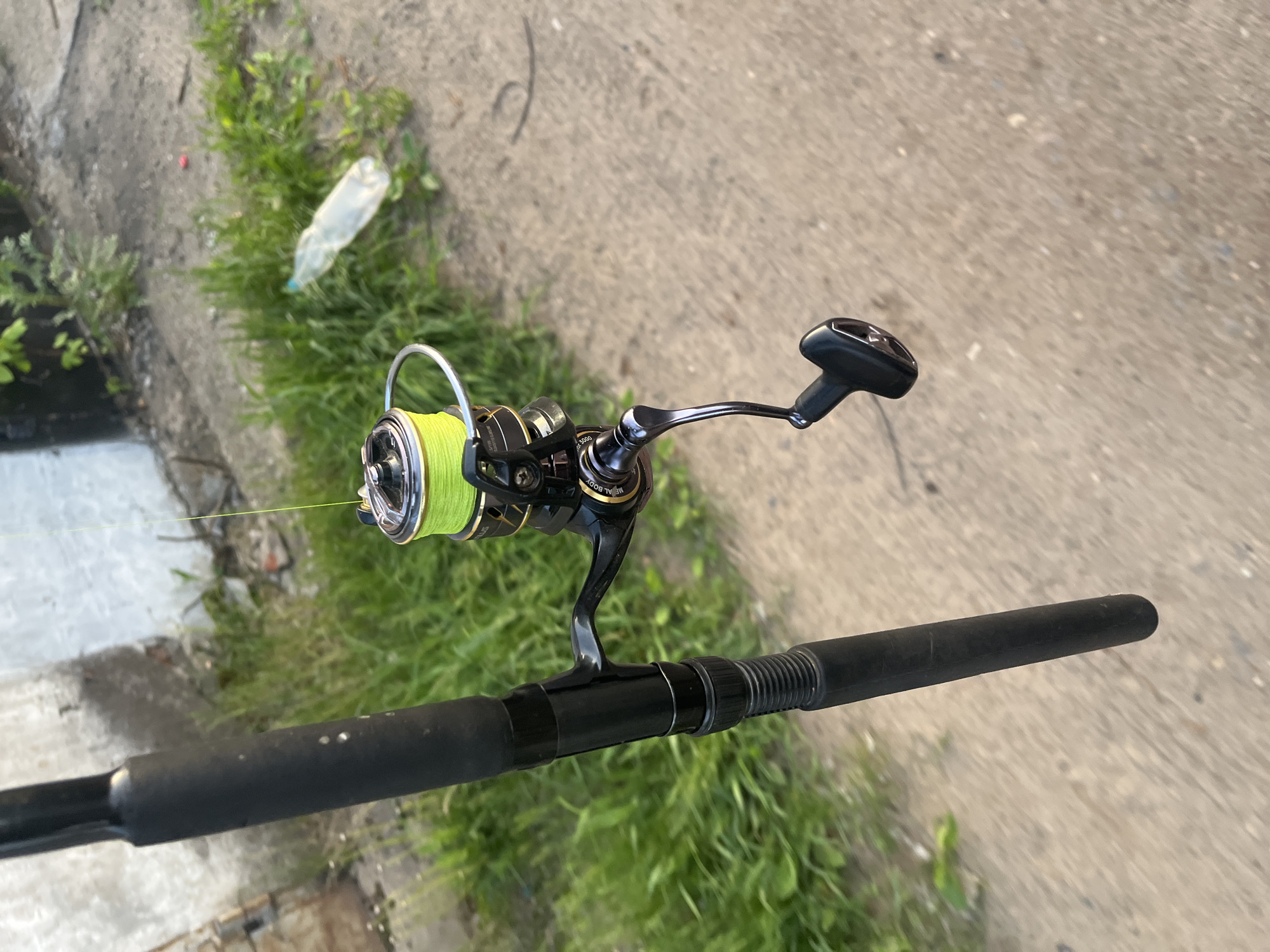 A close-up, eye-level shot captures a fishing rod with a reel, set outdoors during the daytime. The perspective is looking down at the equipment and the ground.
The primary subject is a black fishing rod, visible from its middle section towards the handle. Attached to the rod is a spinning fishing reel, predominantly black and silver, with bright neon yellow fishing line tightly spooled around it. The reel's bail arm is in the open position. Its handle, also black with a silver arm, extends to the right. Visible text on the reel includes "METAL BODY", "3600", and "4000", likely indicating its construction and size. The rod's handle grip, made of a dark, textured material, is visible on the far right and left of the reel assembly.
The background consists of a light grey concrete or paved surface, bordered by a strip of vibrant green grass and weeds. In the grassy area, a clear plastic bottle lies discarded, along with a small, unidentifiable red object. The lighting is bright and even, suggesting a clear day.
The setup of the fishing rod and reel implies that fishing is either in progress, about to begin, or has recently concluded. The scene appears to be an outdoor location with both man-made and natural elements, possibly along a pathway or embankment. The location context for this media file is Chaikovsky, Russia.