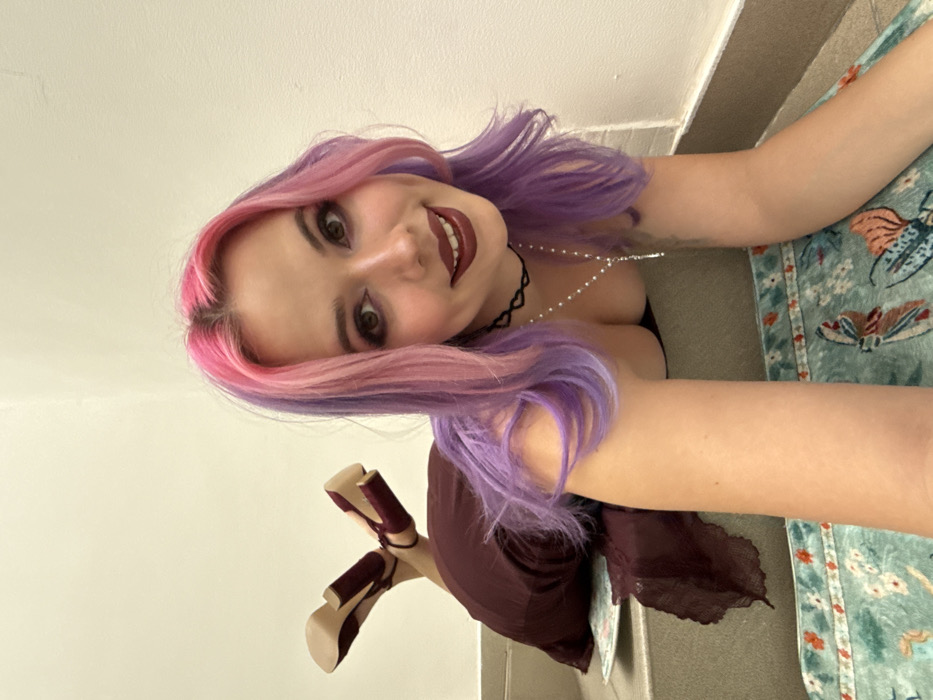 The image displays a young woman lying on her back, looking directly at the camera from a low angle. The image is rotated, making it appear as if she is lying on a vertical surface.
The woman has striking, two-toned hair, starting with bright pink roots that transition into vibrant purple along the mid-lengths and ends. Her face is framed by her hair, and she has a broad, cheerful smile, showing her teeth. Her eyes are accentuated with dark eyeliner and eyeshadow, and her eyebrows are well-defined. She is wearing dark, possibly burgundy or deep red, lipstick.
Around her neck, she wears a black choker with a small, heart-shaped detail at the front. A delicate, thin necklace with small white beads or pearls drapes down her chest, partially visible over a dark-colored top that reveals some cleavage. Her arms are extended to her sides, resting on a patterned surface.
Her legs are slightly bent, and her feet are crossed at the ankles. She is wearing stylish high heels with thick, block heels and ankle straps, in a color that matches her lipstick, a deep red or burgundy. Part of a dark, flowing garment, possibly a skirt or dress, is visible around her legs.
The background on the left side of the image (which would be above her head if the image were upright) is a plain, light-colored wall, likely off-white or light beige. On the right side (where her body and arms rest), there is a patterned fabric or rug. This fabric features a light teal or blue-green background adorned with colorful motifs, including what appear to be butterflies or stylized patterns in shades of orange, yellow, and brown. A distinct line, possibly the base of a wall or a floor edge, runs along the bottom and right side of the frame.
The overall impression is one of playfulness and happiness, with the woman engaging directly with the viewer through her smile and gaze. There is no visible text in the image.