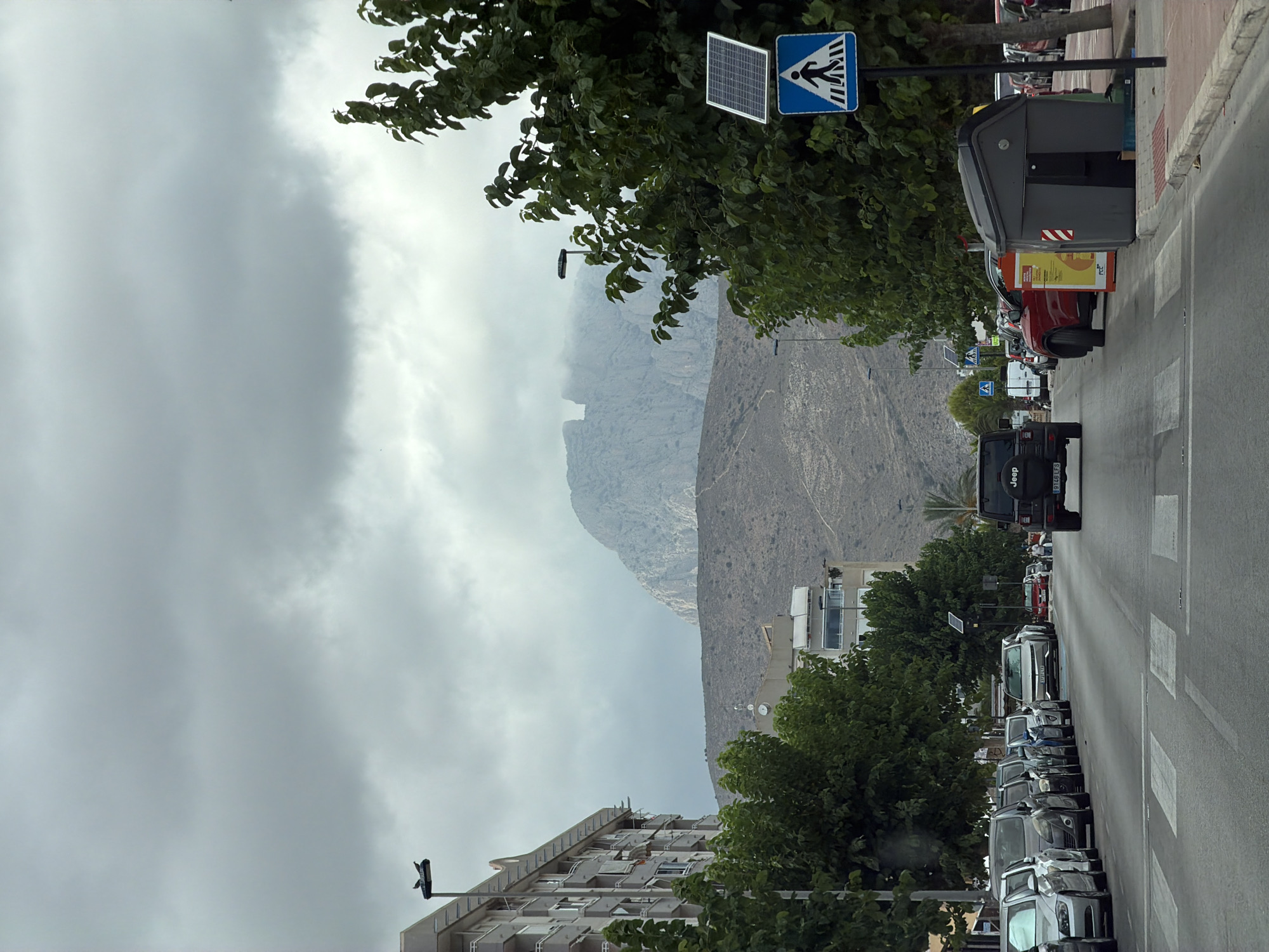This media file depicts a street scene in La Vila Joiosa, Spain, under an overcast sky. The perspective is tilted, as if the camera is held vertically. In the foreground, a line of parked cars stretches along the right side of the road, with a pedestrian crossing visible. The vehicles appear to be a mix of modern sedans and SUVs, with one prominent black Jeep visible closer to the bottom of the frame, bearing the text "Jeep" on its rear.
A large, dark grey trash receptacle sits on the sidewalk to the left of the road, with some orange and yellow signage on its side. Further down the street, trees with lush green foliage line the sidewalk, partially obscuring a building. Behind the trees and buildings, a rugged, mountainous landscape rises, partially shrouded in mist or clouds, suggesting a possibly humid or cloudy day.
On the left side of the frame, a building with multiple balconies is visible, also surrounded by trees. A street light with two bird-like figures perched on it is also present.
Above the street and trees, a blue sign with a white pedestrian symbol is mounted on a pole, accompanied by a small solar panel. The overall atmosphere is subdued, likely due to the cloudy weather. The image appears to capture a typical day in a coastal town with a backdrop of natural elevation. No people are clearly visible in the scene. The time of day is not explicitly indicated, but the diffused light suggests it is daytime.