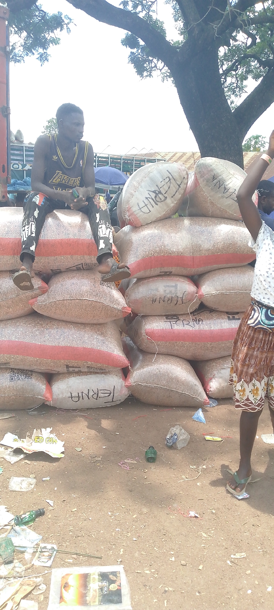 The image depicts a street scene in Tor-Donga, Nigeria, with a focus on a man sitting atop large sacks of grain. He is wearing a dark sleeveless shirt with "LAKERS" across the chest and black pants adorned with white designs. His feet are bare, clad in sandals. The sacks are piled high and appear to be made of a translucent material, some with red banding. The word "TERNA" is written in black marker on several of the sacks. In the background, a large tree with prominent branches extends across the upper portion of the frame, and a bright sky is visible. To the right, a portion of a person's legs and patterned skirt is visible, with their arm raised. The ground is dirt and littered with various debris, including plastic bags, crumpled paper, and a green object. A partially visible sign with the words "ED GIN" is present on the ground in the lower left. The overall impression is of an outdoor market or storage area.