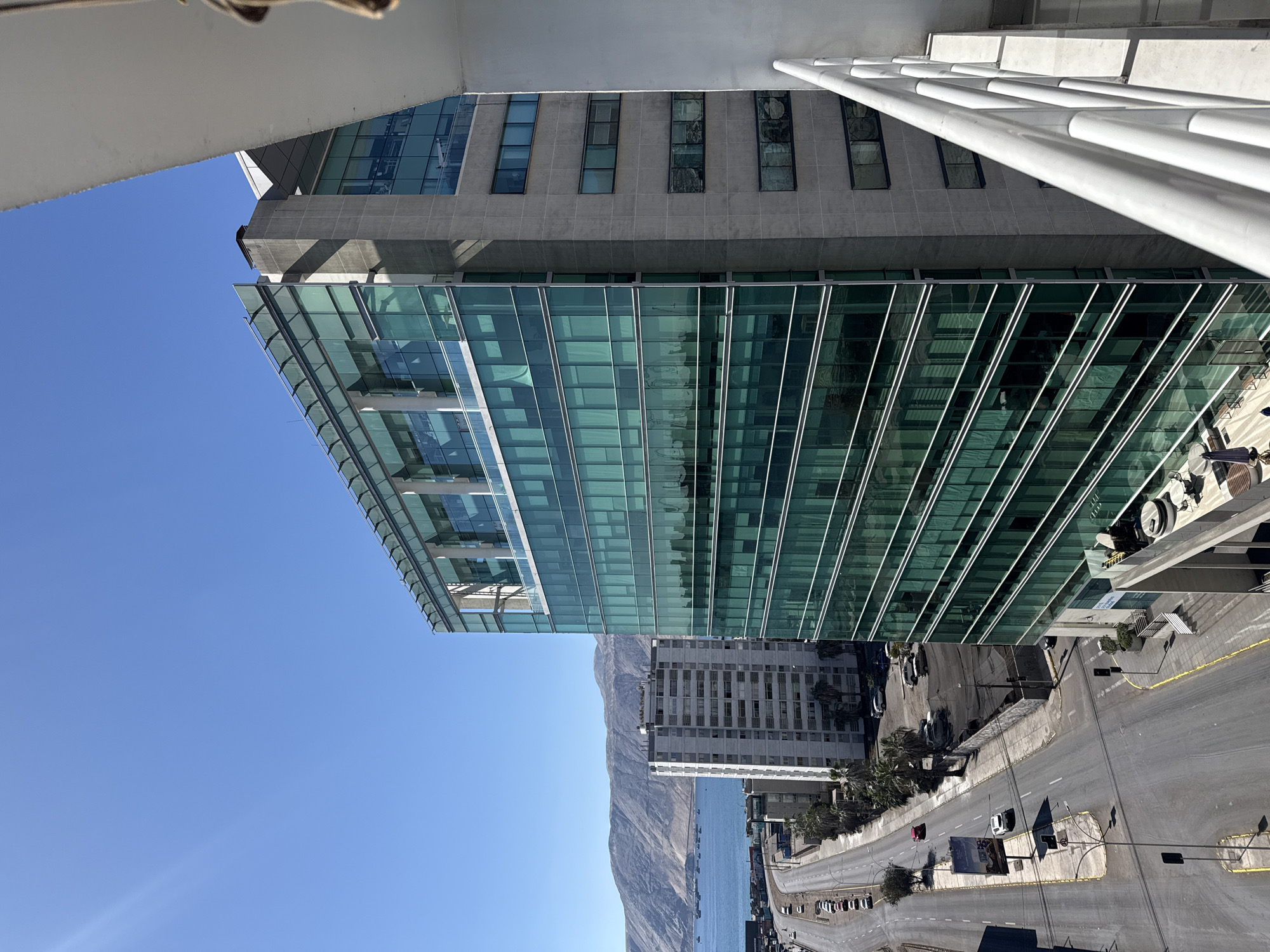 The image, taken from an elevated vantage point in Iquique, Chile, captures a view of a modern glass-fronted office building situated near the coastline. The building, a prominent structure with a teal-tinted glass facade, stands tall against a clear blue sky. To its left, a multi-lane road runs parallel to the coast, lined with parked cars and some palm trees. Across the road, a long, white residential building is visible, and beyond that, the blue waters of the ocean stretch out towards a distant, arid mountain range. Several cargo ships are anchored in the bay. In the foreground, a white railing is partially visible, suggesting the photo was taken from a balcony or similar elevated structure. The scene appears quiet and orderly, with no people actively engaged in activity visible in the immediate vicinity. The sun is shining brightly, casting shadows on the road and buildings.