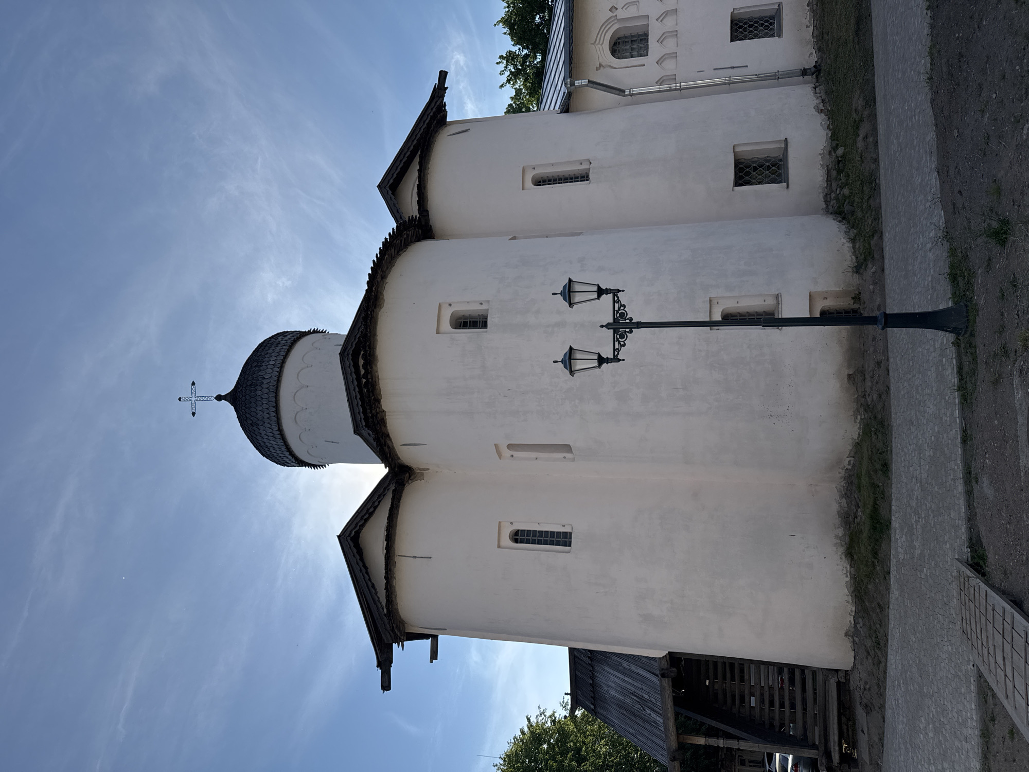 The image shows the Church of the Transfiguration of the Savior on Ilyina Street in Veliky Novgorod, Russia. The white, domed church has a distinct architectural style with multiple rounded protrusions and steeply pitched roofs. A black lamppost with two lanterns stands in front of the church. The sky is blue with some wispy clouds, and the sun is visible behind the dome, creating a bright halo. The ground in front of the church is paved with stone, and a wooden walkway extends into the foreground. There are trees and a part of another building visible in the background. The scene is captured from a low angle, emphasizing the grandeur of the church.