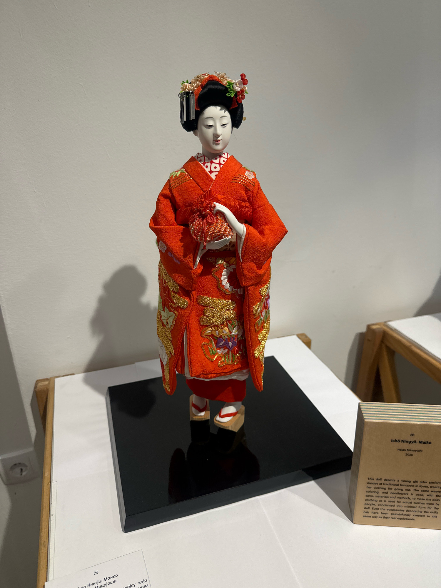 An Izoh Ningyō doll, depicting a Maiko, is displayed on a black, reflective base. The doll is dressed in a vibrant orange kimono with intricate gold embroidery featuring floral motifs. The Maiko has a serene expression, traditional Japanese hairstyle adorned with flowers, and is holding a small, beaded red pouch. The doll is positioned on a white surface, casting a shadow on a pale grey wall behind it. To the right of the doll is a small wooden information stand with text describing the doll. The background consists of a plain wall and the corner of a wooden structure, suggesting an indoor exhibition or gallery setting. The overall lighting is soft, illuminating the doll and its details. The location is identified as the Museum of Yugoslavia in Belgrade, Serbia.