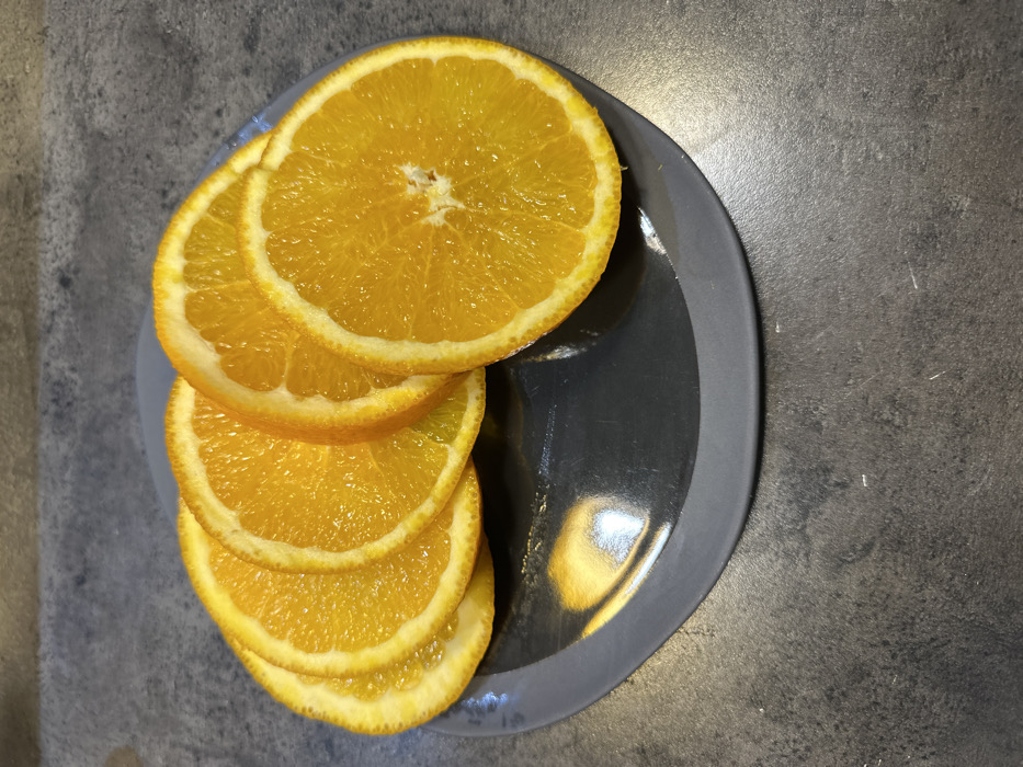 Six circular slices of orange are displayed on a dark grey ceramic plate. The slices, approximately 0.5 cm thick, exhibit a vibrant orange color of the pulp and a thin white pith rim. Five slices are fanned in an overlapping arrangement, curving from the left to the center of the plate, with the sixth prominent slice positioned on the right. The plate rests on a dark grey, mottled surface, consistent with a stone countertop or table. The background comprises the same dark, textured surface. No individuals, specific actions, or interactions are present. The location of the scene, specifically whether it is in Tbilisi, Georgia, cannot be ascertained from the visual evidence provided.