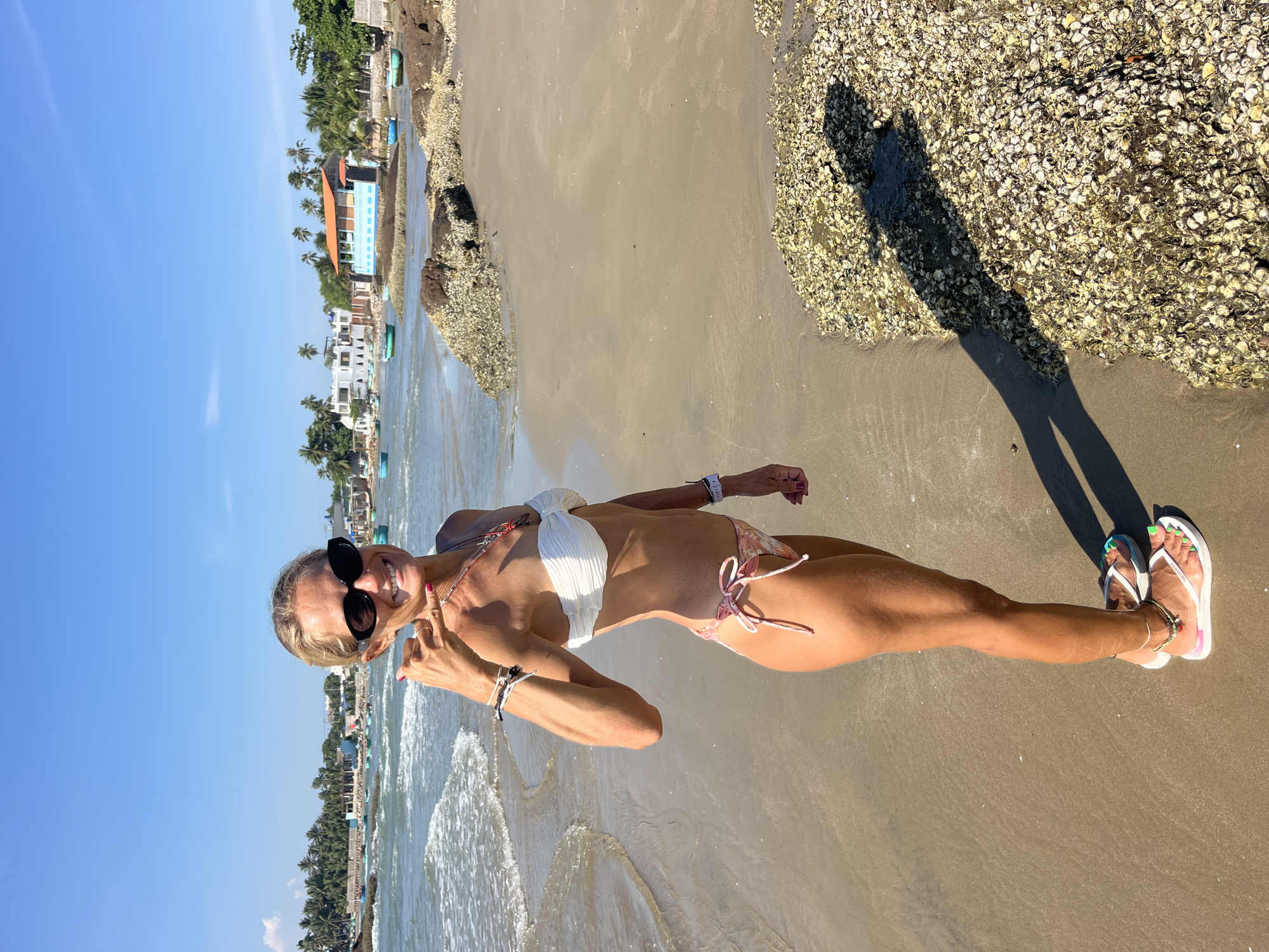 In Mũi Né, Vietnam, a woman stands on a sandy beach under a clear blue sky. She is wearing a white strapless bikini top and a floral-patterned bikini bottom. She has dark sunglasses on and is smiling at the camera, holding her hand up in a "shaka" gesture. Her toenails are painted bright green, and she wears a few necklaces and bracelets. The ocean waves gently lap at the shore behind her, and in the distance, small boats are visible. The beach is dotted with rocks covered in shells. A shadow of the woman is cast on the sand in the foreground.