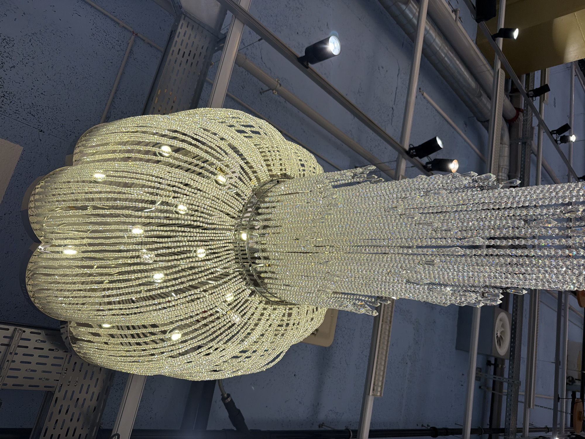 A low-angle, indoor shot shows a large, decorative chandelier hanging from the ceiling. The chandelier consists of numerous strands of beads, arranged in a rounded, flower-like shape at the top and a more rectangular shape below. The strands of beads are illuminated, and light is reflected off of them, creating a sparkling effect. The background reveals a network of metal beams, pipes, and electrical conduits attached to a light blue ceiling. Several track lights are also mounted on the beams, pointed in different directions. The presence of the industrial-style ceiling suggests that the location might be in a commercial or industrial space. There are no people visible in the frame. It is impossible to determine the exact location within Spreitenbach, Switzerland, based solely on the image.
