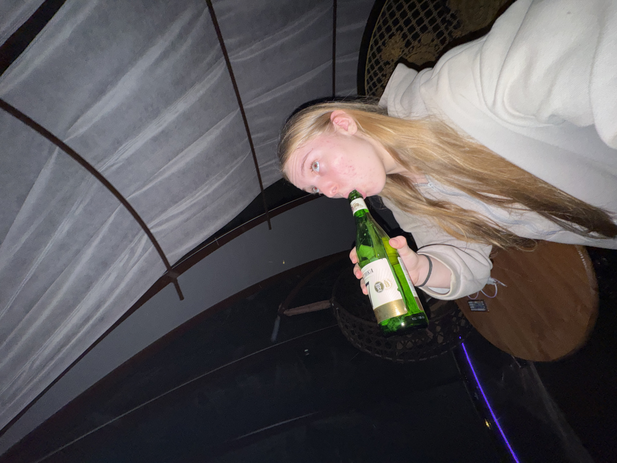 A blonde woman in a light-colored hoodie is drinking directly from a green glass bottle. She is indoors, beneath a white, draped structure, possibly netting. A round, wooden table is visible, along with a woven seat. The bottle has a label with Russian text, and a partial word reads "IKA." The scene is dimly lit. While the presence of Russian text on the bottle label suggests a location in Russia, the specific city of Zhigalki cannot be determined from the image alone.