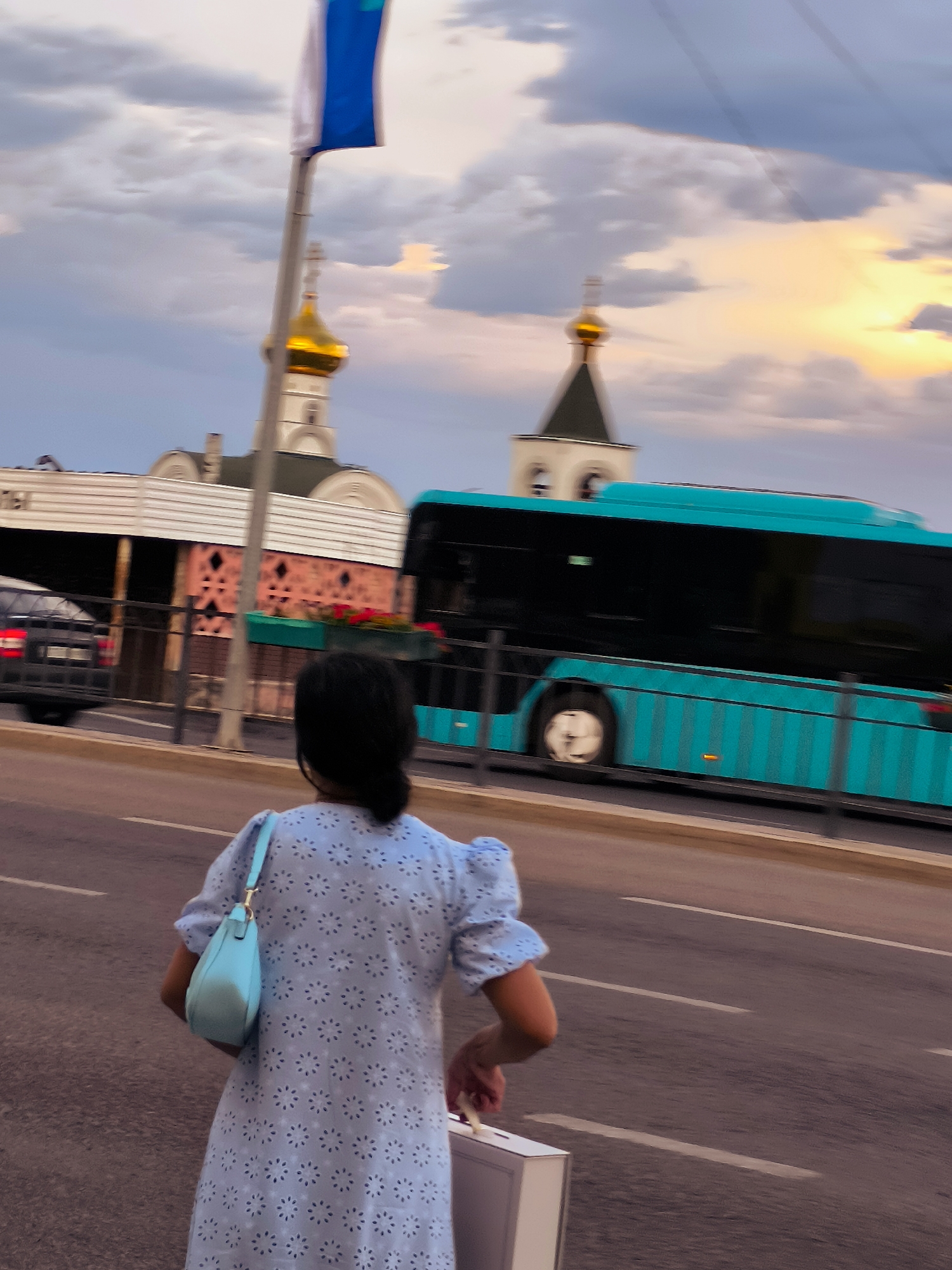 A young woman with dark hair is shown from behind, walking on a street with a light blue patterned dress and a pale blue purse. She is carrying a white rectangular object in her right hand. In the background, a bright teal-colored bus is in motion on a road. Behind the bus, a church with a golden dome and a white steeple is visible, along with another building that appears to be part of a civic or religious complex. A tall flagpole with a blue and white flag is also present. The sky is partly cloudy with a warm sunset glow. The image captures a street scene in Qarağandı city administration, Kazakhstan.