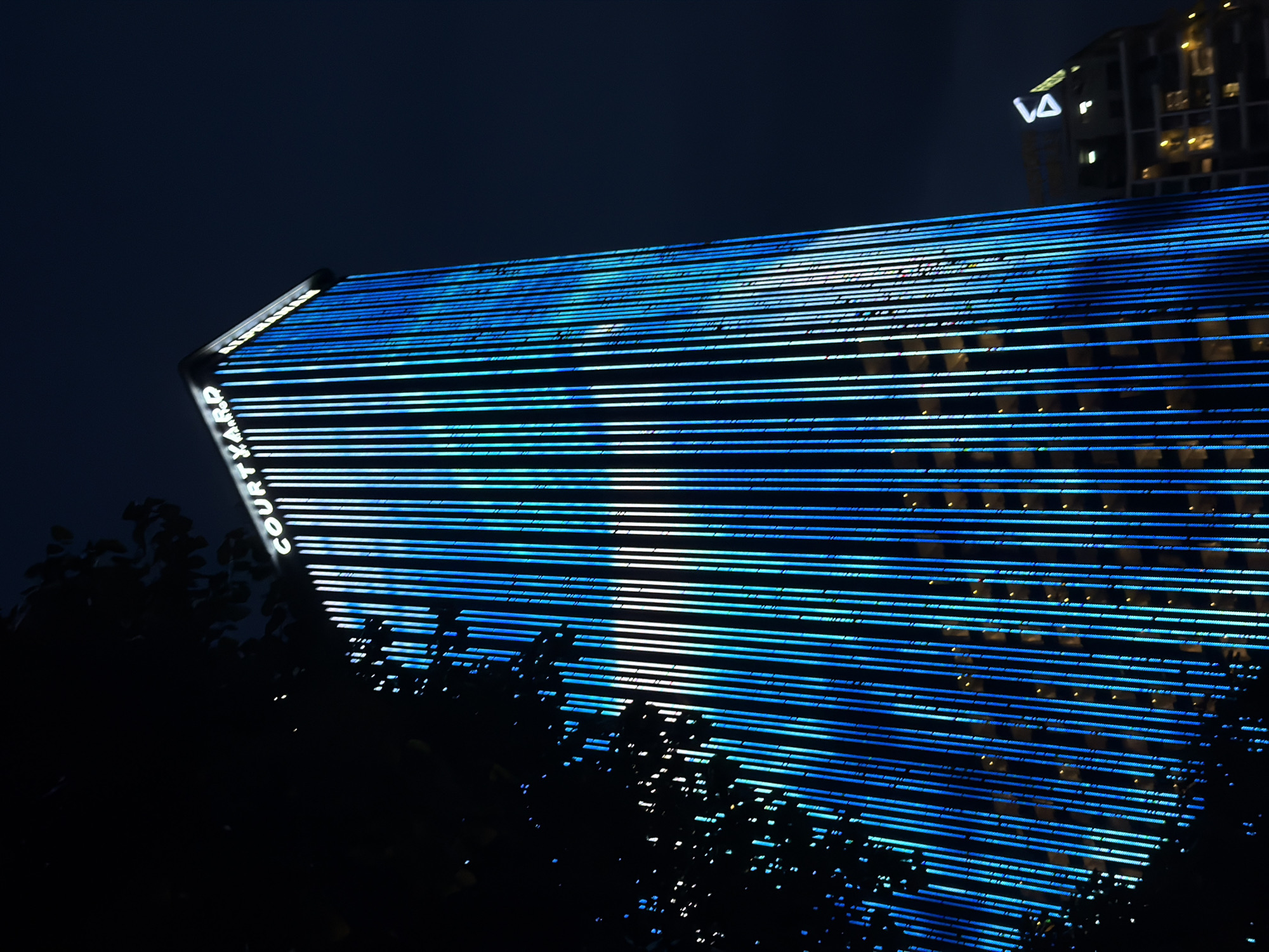 An exterior, low-angle, eye-level shot captures a skyscraper adorned with vertical blue lights at night in Da Nang, Vietnam. The building's facade is illuminated by numerous thin, glowing blue lines that run from top to bottom, creating a striking visual effect against the dark sky. The word "COURTYARD" is visible in illuminated white letters across the top of the building. To the right of the main skyscraper, another building with a modern, angular design is partially visible, also illuminated by interior lights and a distinctive triangular logo. A dark silhouette of a tree with branches and leaves partially obscures the left side of the main building, adding depth and contrast to the scene. The overall atmosphere is modern and urban, with the vibrant lights of the skyscraper dominating the composition.
