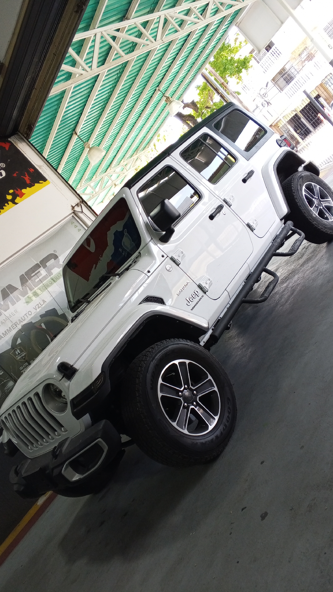 The image depicts a white, four-door Jeep Wrangler, likely parked inside an automotive service center, garage, or showroom. The vehicle is positioned at an angle, prominently showcasing its front and side profiles. The Jeep features a classic seven-slot grille, round headlights, black bumpers with integrated fog lights, and black side mirrors. It is equipped with multi-spoke alloy wheels, shod with Goodyear tires, and has robust black side steps or rock sliders. The "Jeep," "Wrangler," and "SAHARA" badges are visible on its side. Reflections on the windshield show a multi-colored pattern, possibly from a flag or another sign.
The setting is an indoor space with an industrial aesthetic. The ceiling features exposed white metal trusses supporting green corrugated metal panels, from which two white light fixtures hang. On the left side of the frame, a large banner or sign is partially visible, displaying text such as "HAMMER," "AUTOMOTRIZ," and "AMMERAUTO_VZLA," suggesting an automotive business located in Venezuela. Other partially visible text includes "H HANSEN." The floor is a dark, possibly concrete or asphalt, surface. Through an opening on the right side of the building, trees and parts of an exterior structure are visible, indicating it is daytime.
No people are present in the image, and the Jeep appears to be stationary, suggesting it is either on display, awaiting service, or parked. The overall lighting is bright, combining natural light from outside with indoor artificial illumination. The scene conveys a clean, functional environment typical of an auto-related establishment.