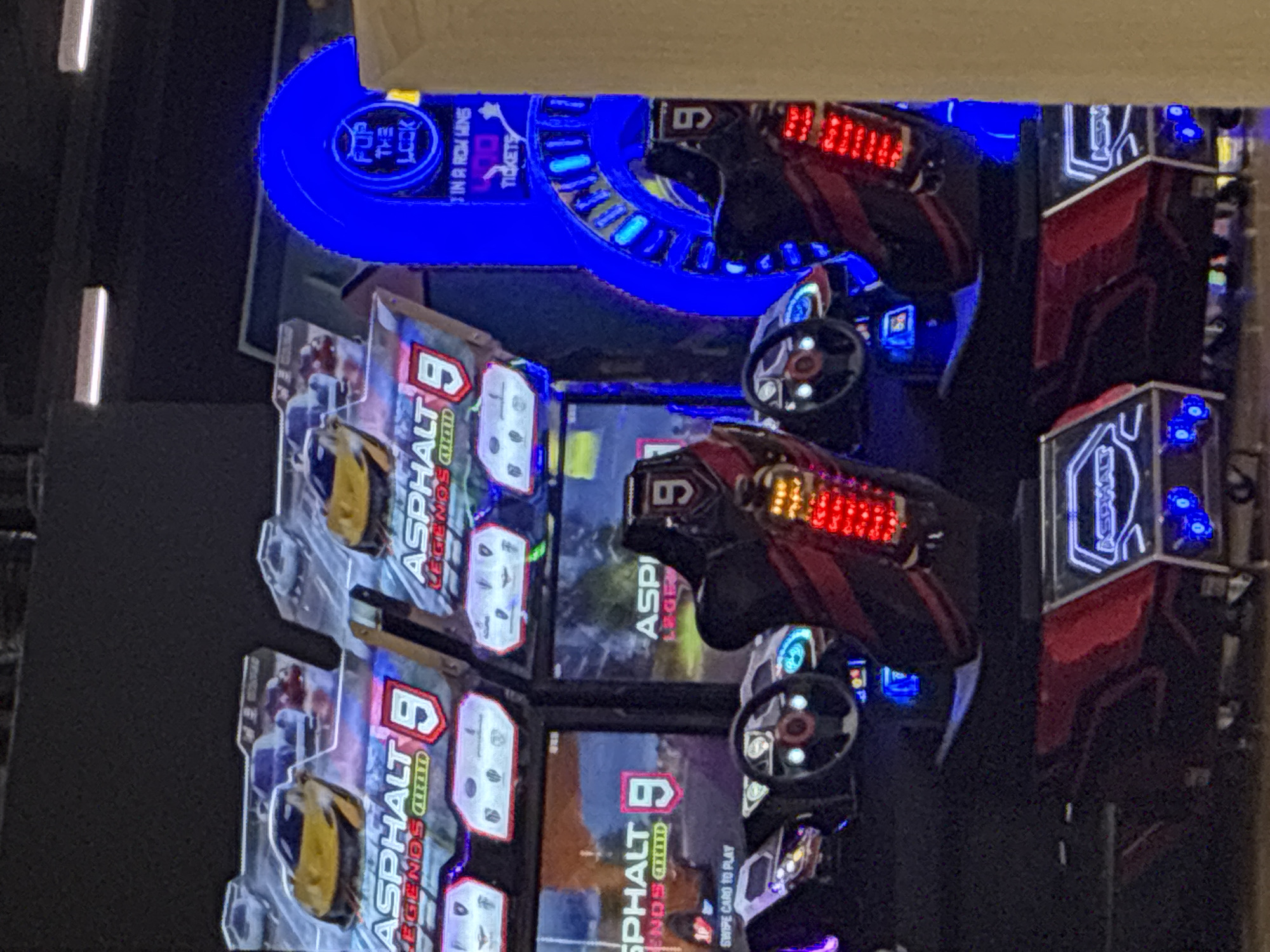 The image shows multiple "Asphalt 9 Legends" arcade racing games, likely in an entertainment venue. Each game has a racing seat, steering wheel, and a large screen displaying the game. The game cabinets are decorated with blue and red lights. Signage indicates a jackpot or bonus ("FUP THE LOCK WIN A ROW WINS TICKETS"). The location is impossible to determine solely from this image, and there is no indication in the image that this scene is in Plovdiv, Bulgaria.