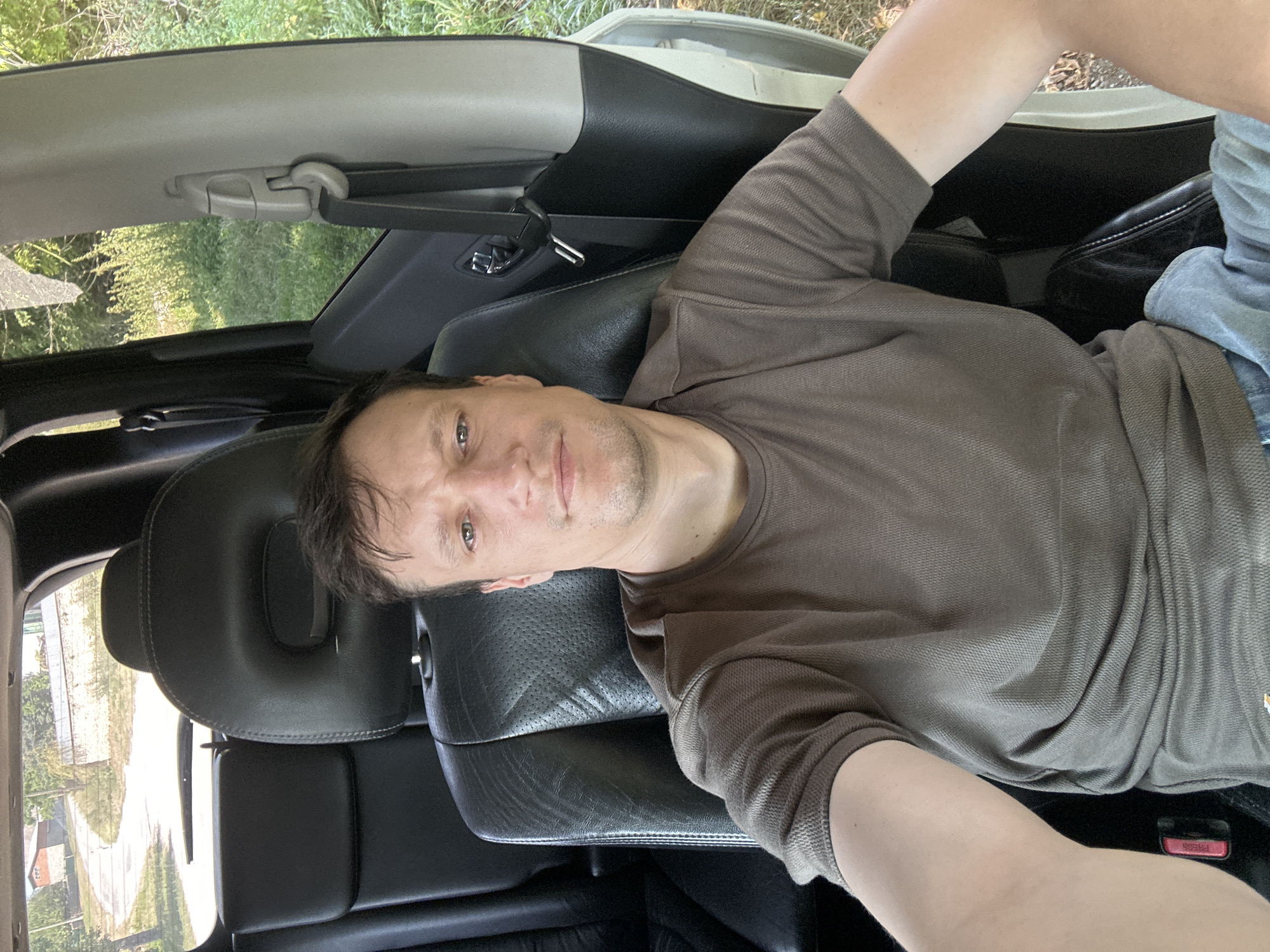A man in his 30s with short brown hair and a light beard is seated in the driver's seat of a car. He is wearing a brown t-shirt and blue jeans. The car's interior is visible, with black leather seats and a seatbelt. The man is looking towards the camera, and his right arm is extended, holding what appears to be a phone, suggesting he is taking a selfie. Outside the car, through the rear window and side window, a rural landscape is visible, with a paved road, buildings, trees, and green vegetation, consistent with the setting of Makiv, Ukraine. The scene is captured during the daytime, with natural lighting.