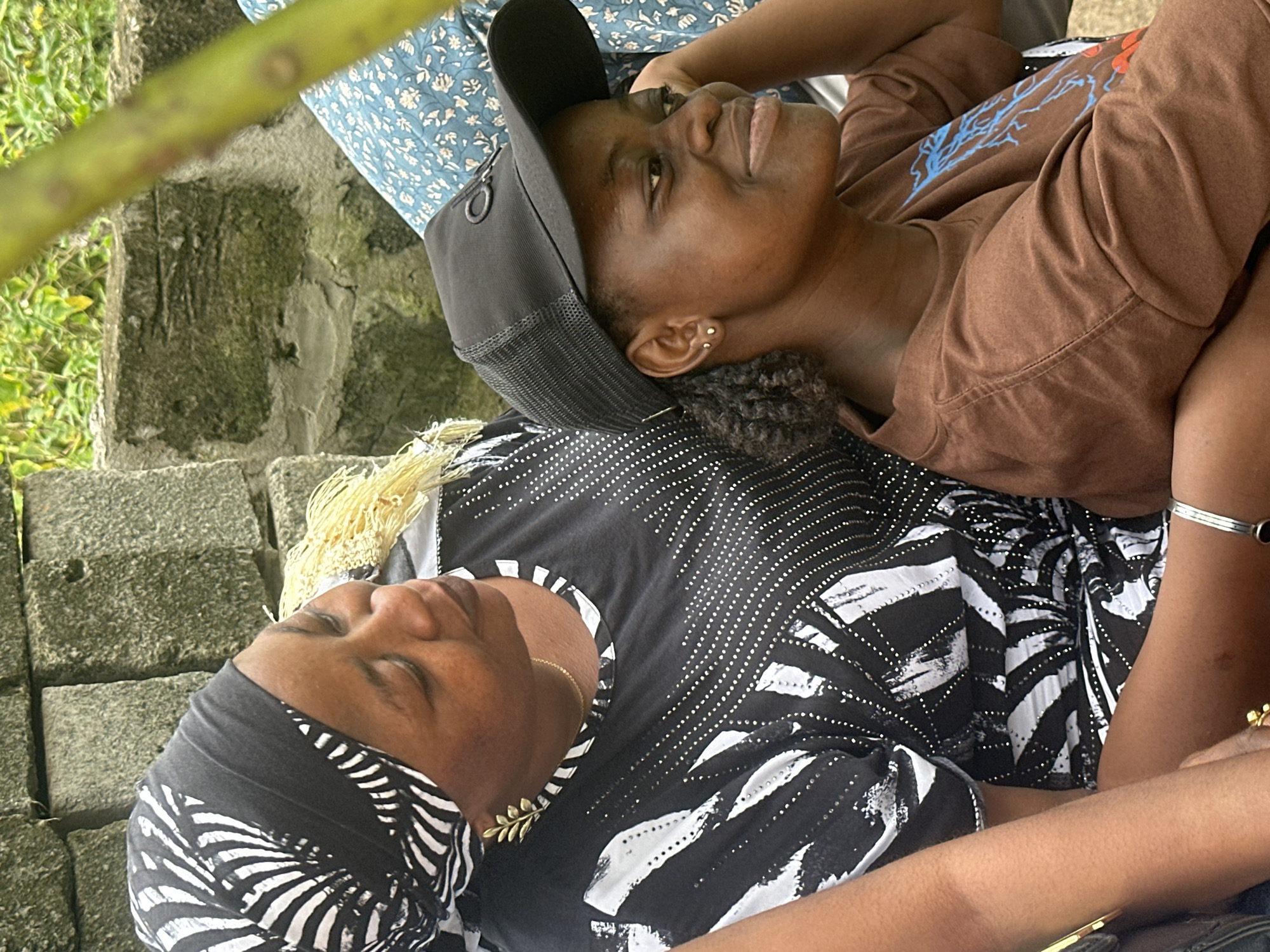 In Ganyingbo, Nigeria, two women are seated outdoors near a brick wall. The woman on the left wears a black and white patterned headscarf and a black top with white patterned accents. She has a leaf-shaped earring. The woman on the right wears a black baseball cap and a brown t-shirt with a graphic design. Both women appear to be looking to their right. The environment includes lush green foliage in the background.
