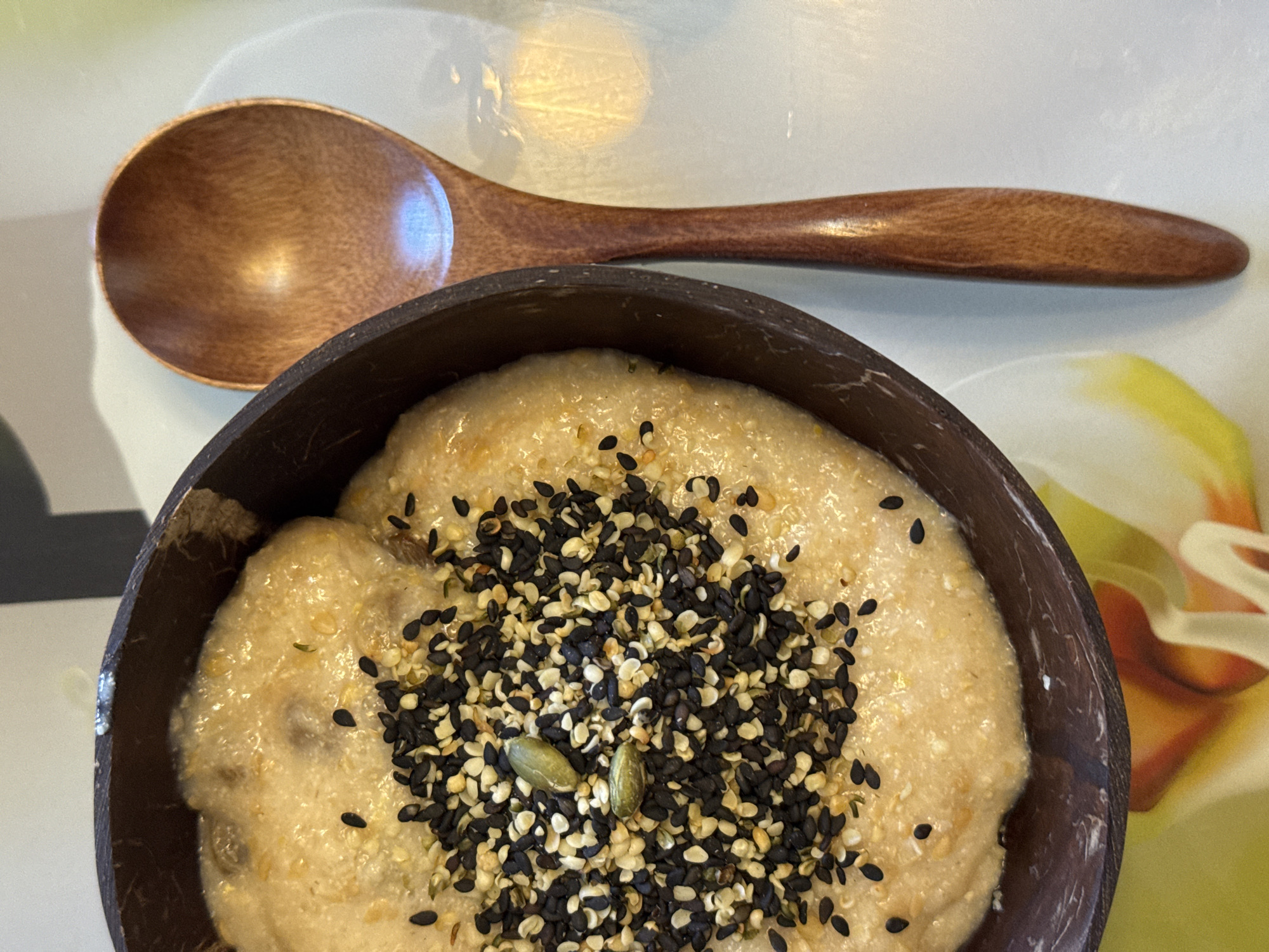 The image shows a bowl of porridge topped with a mix of black and white sesame seeds, as well as a couple of green pumpkin seeds. The bowl appears to be made of dark brown, possibly natural material, perhaps coconut shell. A wooden spoon rests beside the bowl on a light-colored surface. The location is unknown.