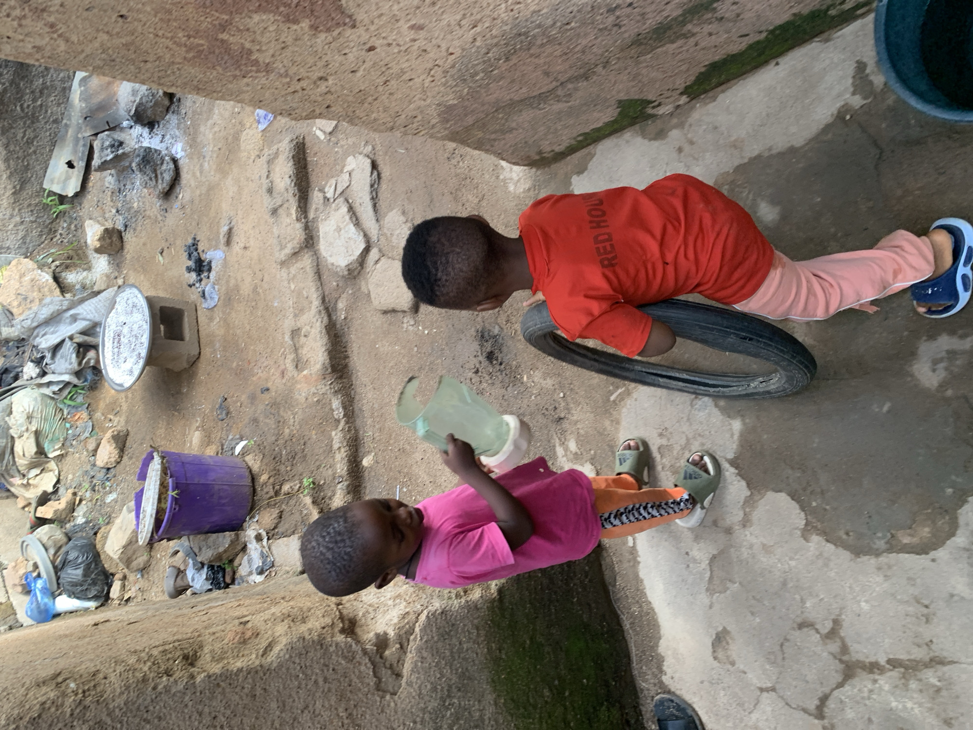 In Jos, Nigeria, two young children are in an outdoor courtyard. The child on the left, facing away from the viewer and to the left, wears a pink t-shirt and orange pants with a black and white stripe pattern, along with green sandals. They are holding a clear plastic pitcher with a pink base. The child on the right, facing away from the viewer and to the right, wears a red t-shirt with "RED HOUS" printed on the back in white and light pink pants, and blue sandals. They are holding a black tire upright. The courtyard is paved with cracked concrete and dirt. In the background, a purple bucket sits near some rocks and debris. Further back, there's evidence of a fire, with scattered ashes and metal sheeting. The environment appears somewhat unkempt, with various discarded items and rough walls suggesting a less developed urban setting.