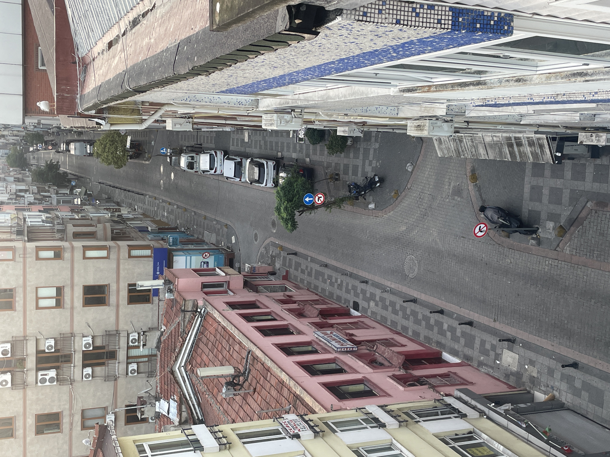 The image depicts a narrow, downward-sloping street in Kadıköy, Turkey, lined with multi-story buildings. The architecture suggests a densely populated urban area with a mix of residential and commercial spaces. On the left, a light yellow building features multiple windows and balconies with air conditioning units. Adjacent to it is a pink building with a red-tiled roof, housing businesses with signage that is partially visible, including one that reads "HAMAMI". The street itself is paved with cobblestones and has a sidewalk on the left.
Several white cars are parked along the street, and a black motorcycle is parked on the sidewalk. Traffic signs are visible, including a "no left turn" sign and an arrow indicating traffic direction. A single seagull is perched on the roof of a building on the right side of the frame, which is covered in metal sheeting and appears to be under repair or construction. The buildings on the right are adorned with blue and white mosaic tiles. The overall impression is a typical street scene in a bustling Turkish city.