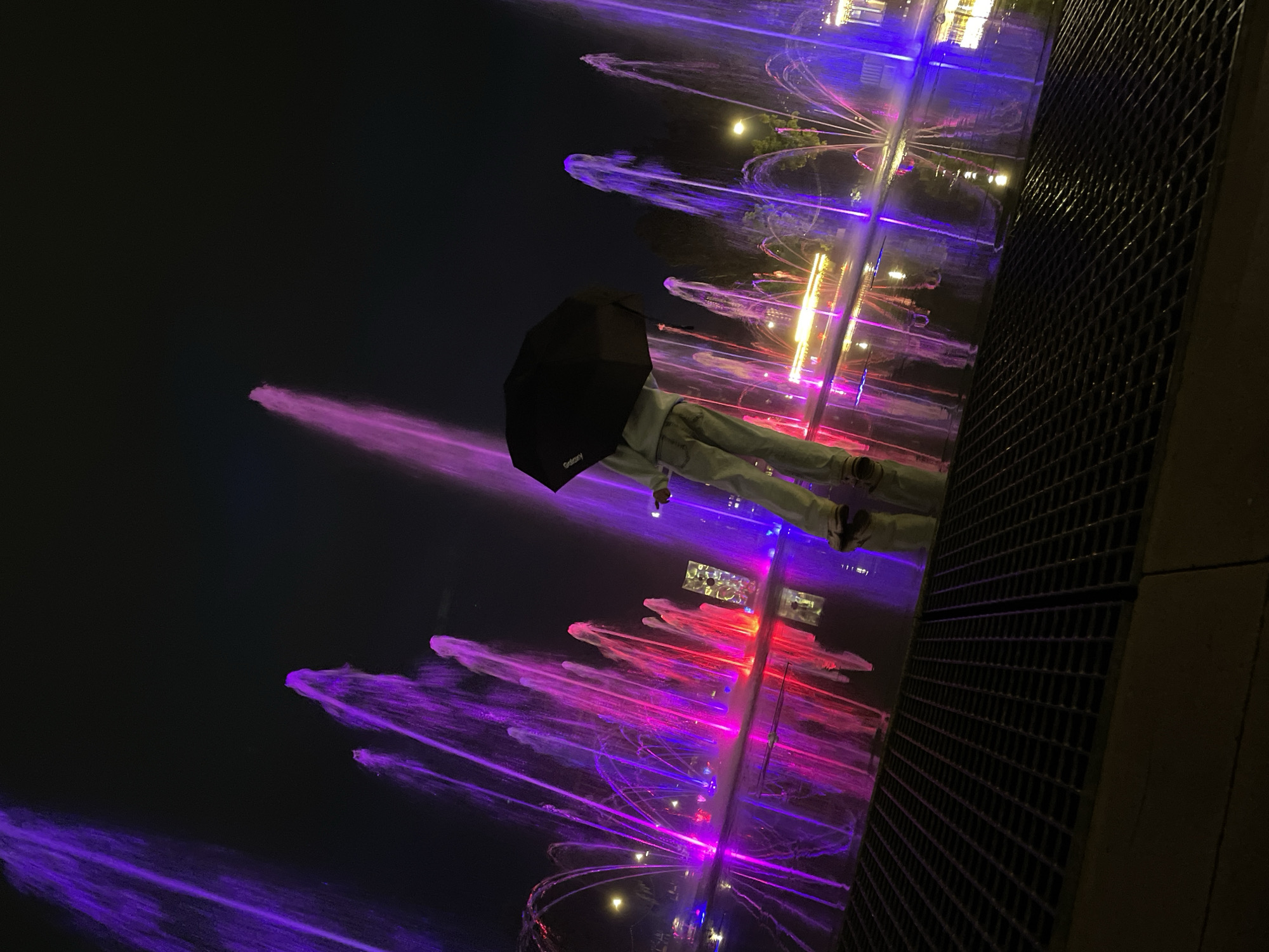 A nighttime shot features a person holding an umbrella in front of a water fountain with colorful illumination in Izhevsk, Russia. The fountain jets shoot water upwards, lit with a mix of purple, pink, and yellow light. The person, dressed in light-colored pants and dark shoes, is positioned such that the umbrella is visible between them and the fountain. Part of a metal grate structure is visible in the foreground. The background is a dark night sky.